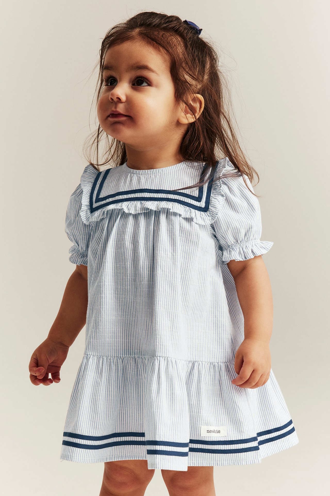 Vorderansicht: Ein kleines Mädchen trägt ein hellblau gestreiftes Baumwoll-Matrosenkleid mit Puffärmeln und Rüschen.