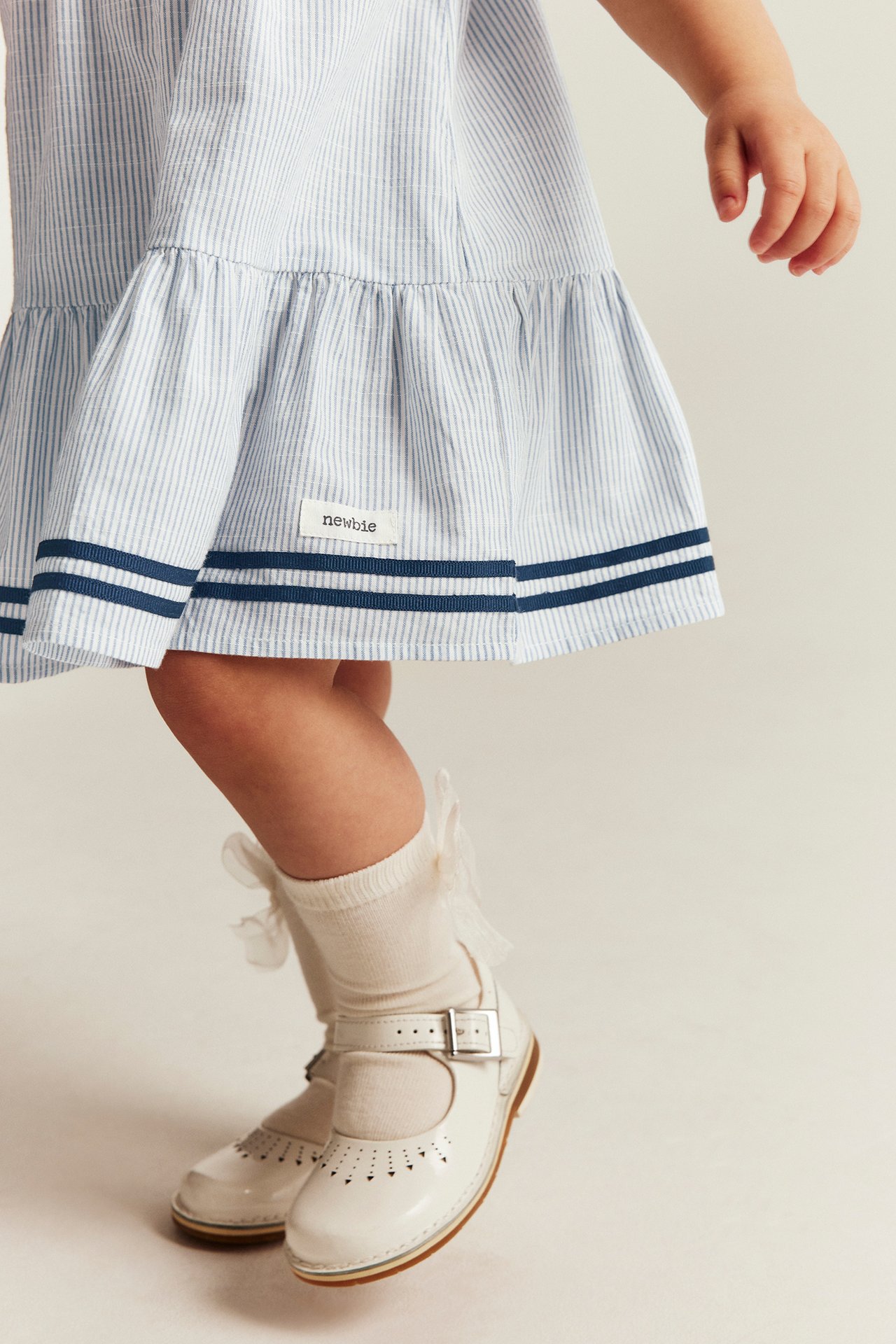 Teilweise Vorderansicht: Baby in blau gestreiftem Baumwollkleid, dunkelblauem Saum, cremefarbenen Socken, weißen Mary Jane Schuhen.