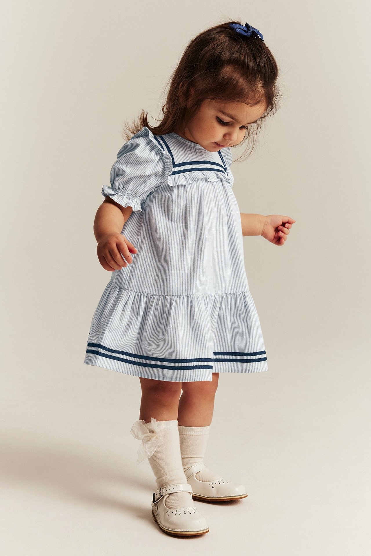 Front view of a kid wearing a blue striped sailor dress with puff sleeves, frills, white socks, and white shoes.