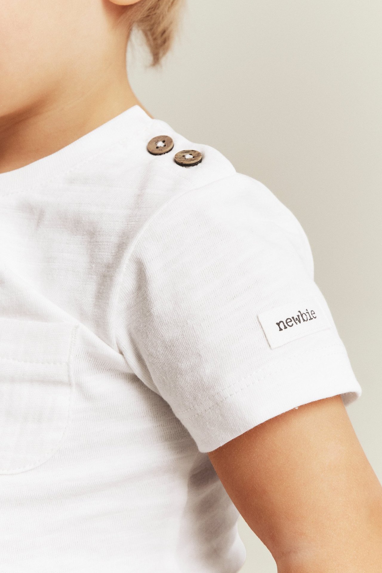 Nahaufnahme eines Babys in einem cremefarbenen, kurzärmeligen Slub-Jersey-Body mit Holzknöpfen und Brusttasche.