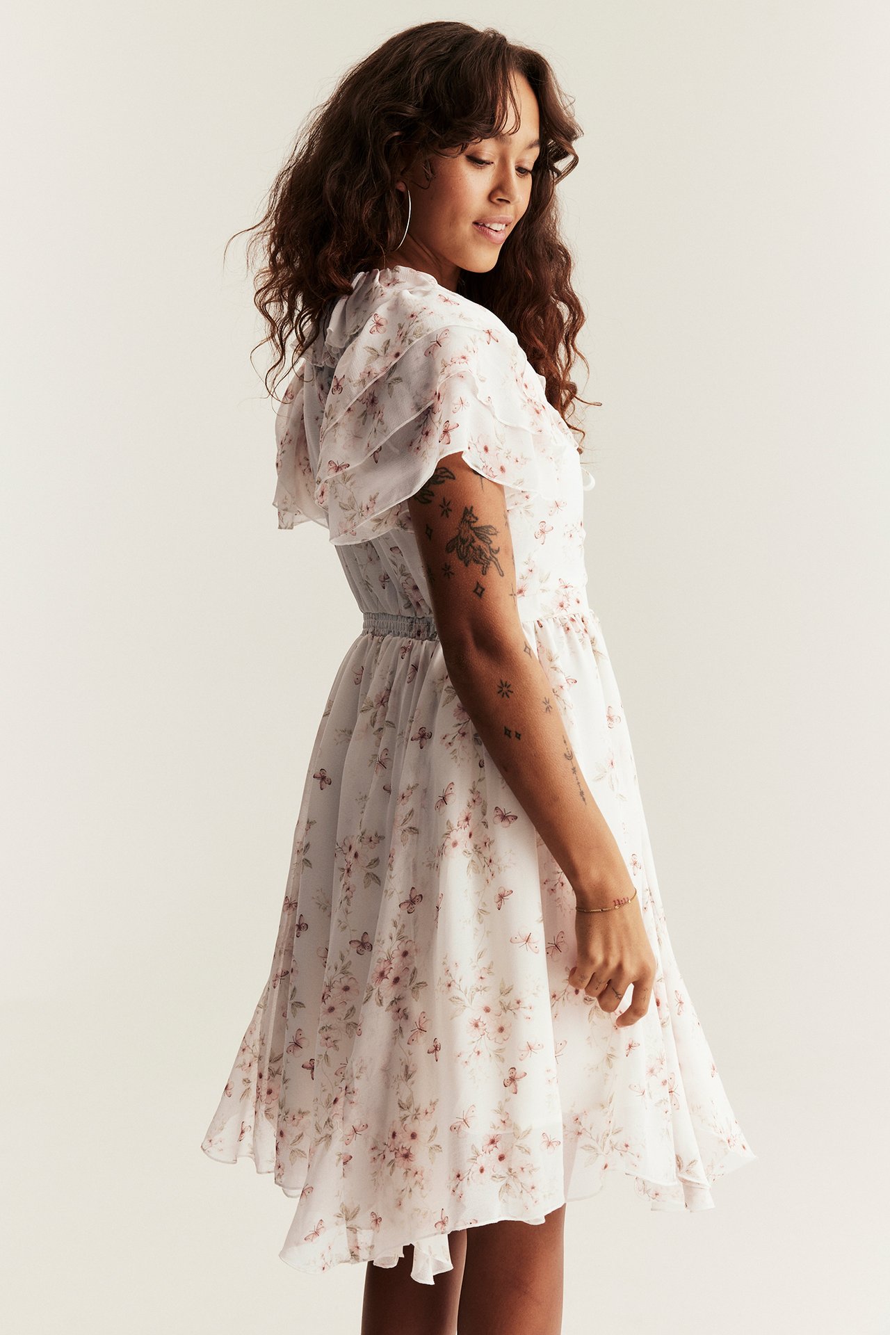 Side view of a woman wearing an off-white floral chiffon dress with ruffle sleeves and an elastic waist.