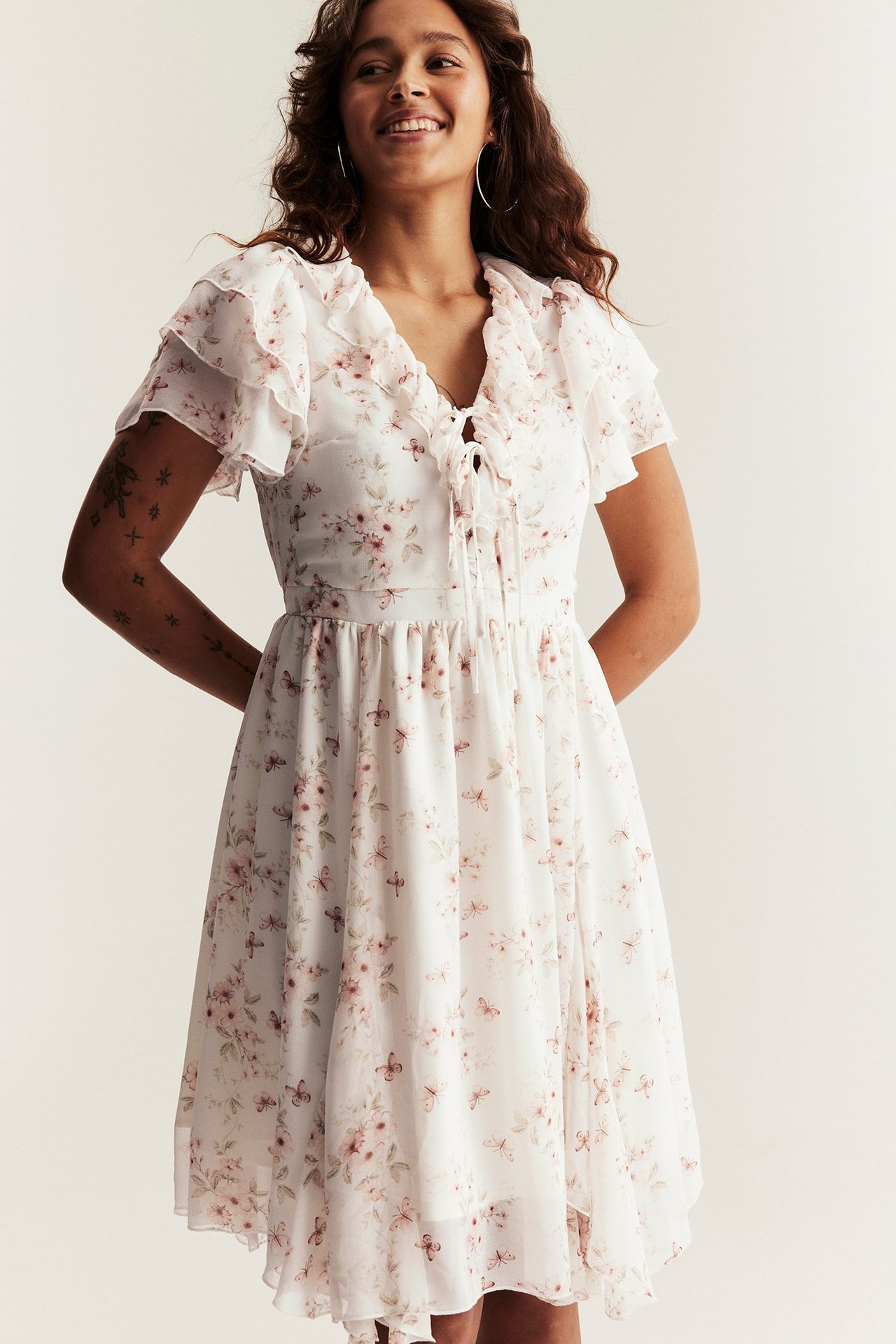 Vorderansicht: Frau in cremeweißem floralem Chiffonkleid mit Rüschen, Bindebändern, kurzen Ärmeln und Creolen.