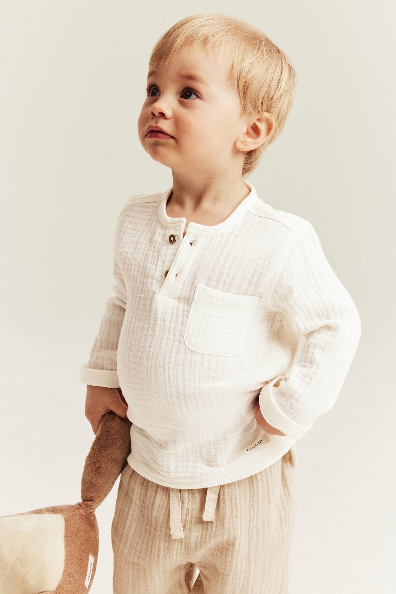 Kind in offwhite langärmeligem Musselin-Oberteil mit Knöpfen, Tasche; beige Hose; hält ein Stofftier.