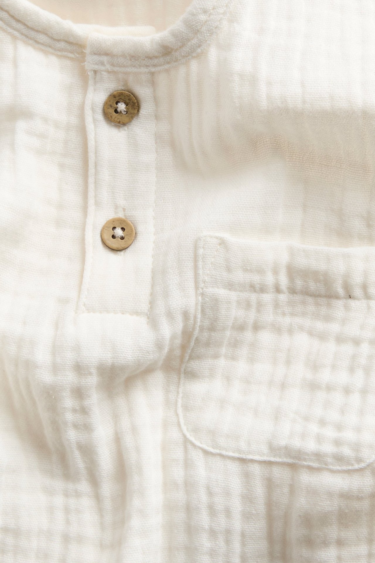 Close-up front view: off-white cotton muslin long-sleeved top with two buttons on placket and a patch pocket.