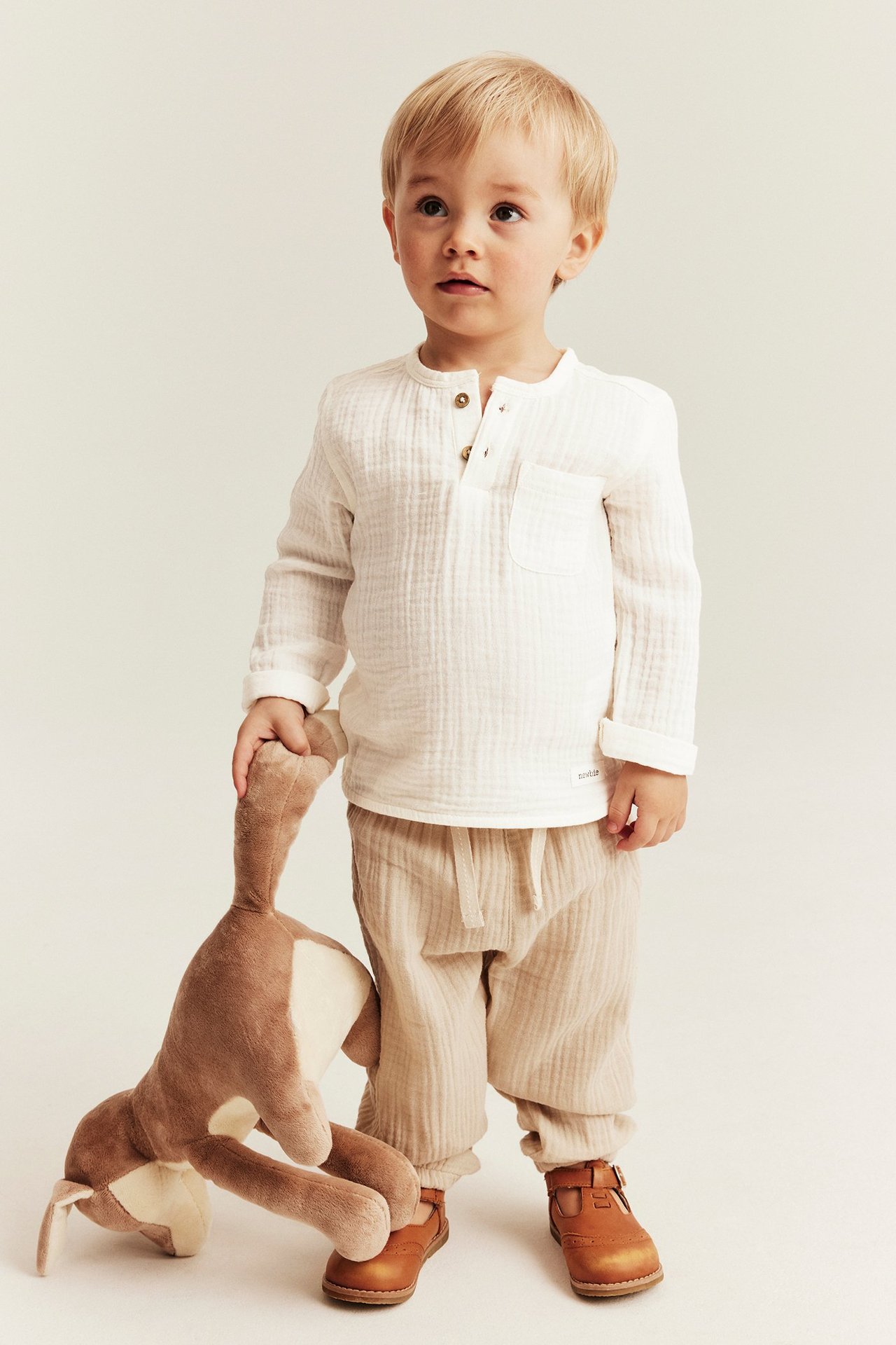 Kind trägt cremeweißen Langarm-Musselin-Top, beigefarbene Hose, braune Schuhe und hält ein Plüschtier.