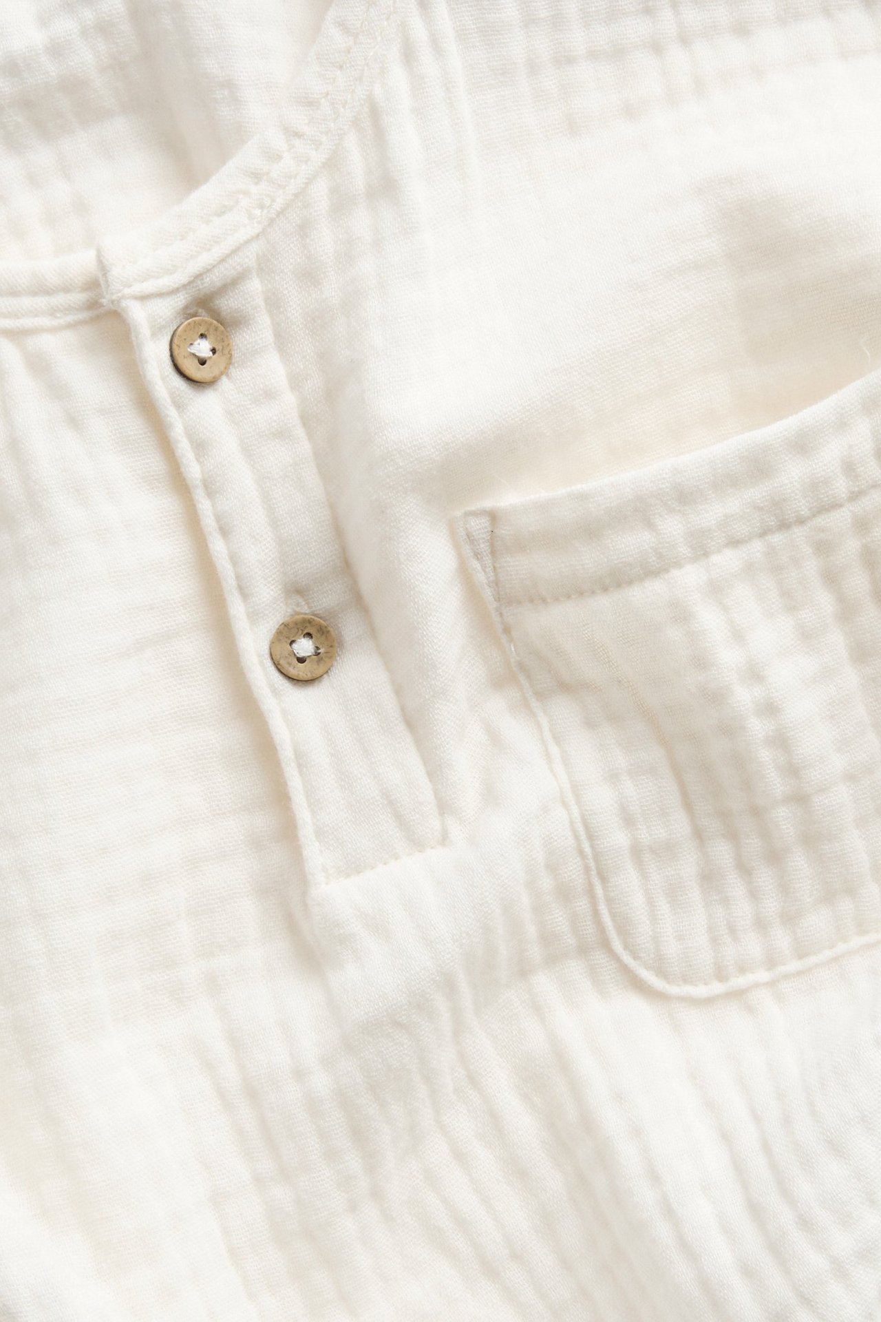 Nahaufnahme eines cremefarbenen langärmeligen Oberteils aus Baumwollmusselin mit zwei Knöpfen und Brusttasche.