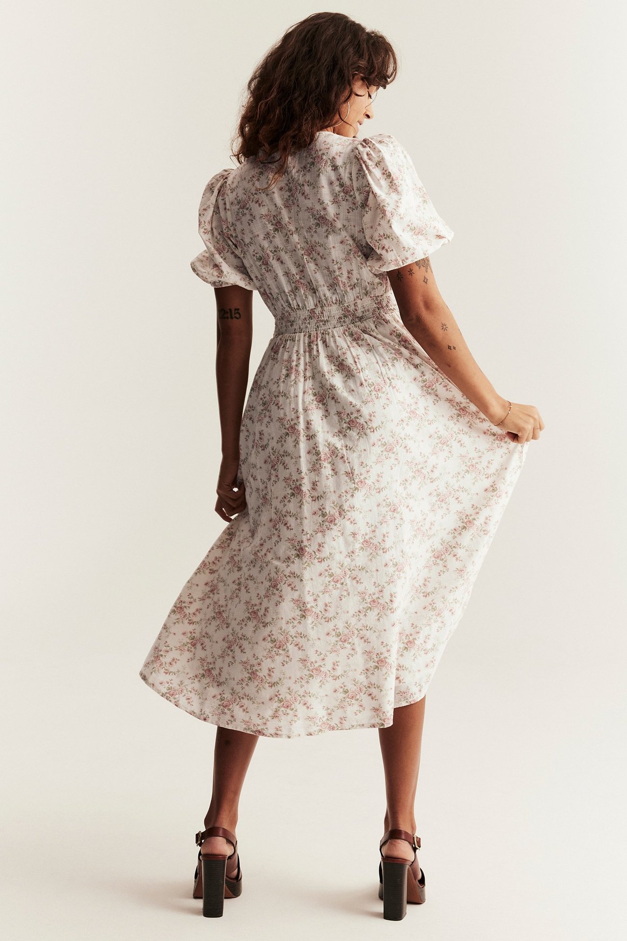 Back view of a woman wearing an off-white floral cotton midi dress with puff sleeves and dark brown block-heel sandals.