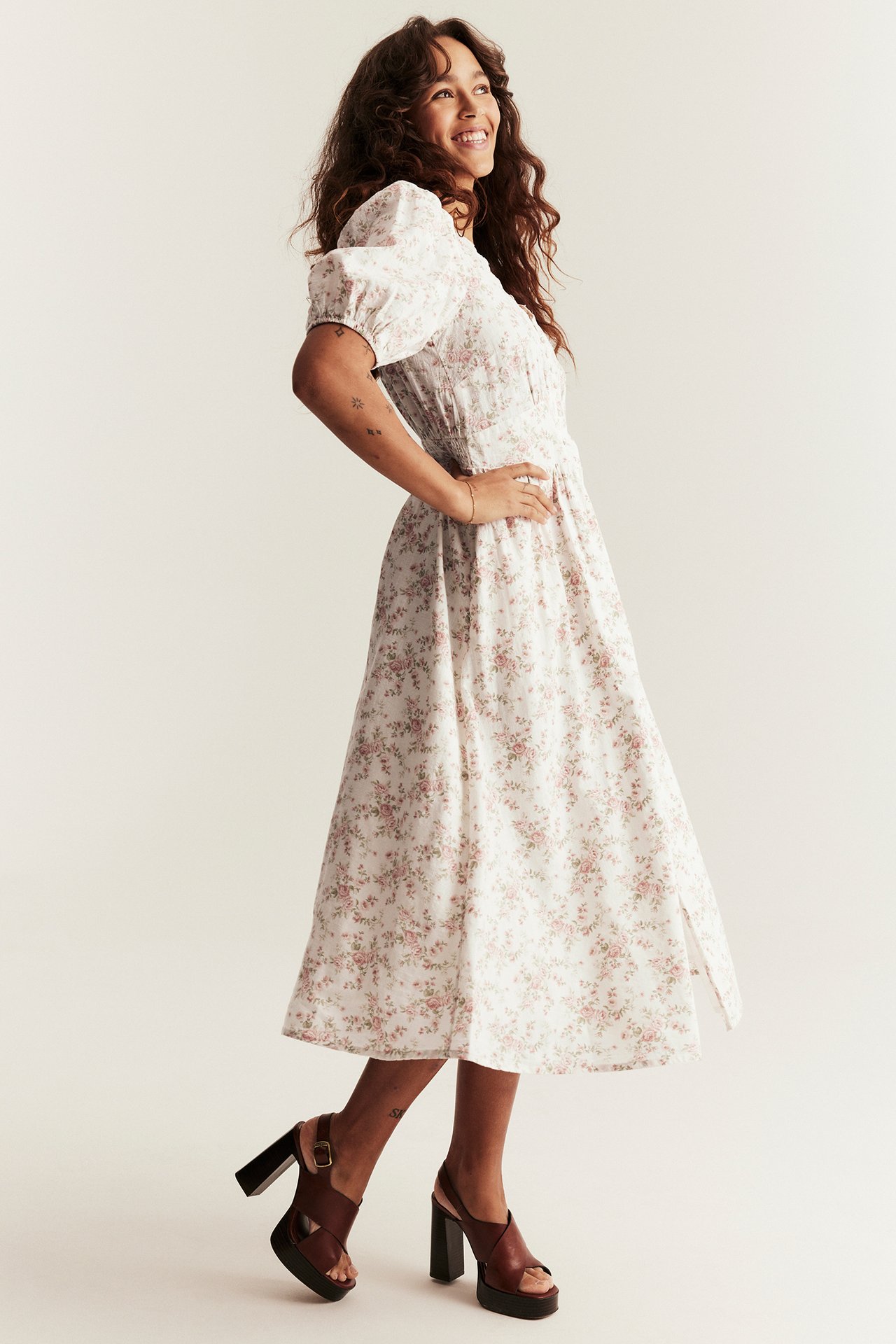 Side view of a woman wearing an off-white floral cotton midi dress with puff sleeves and dark brown platform sandals.
