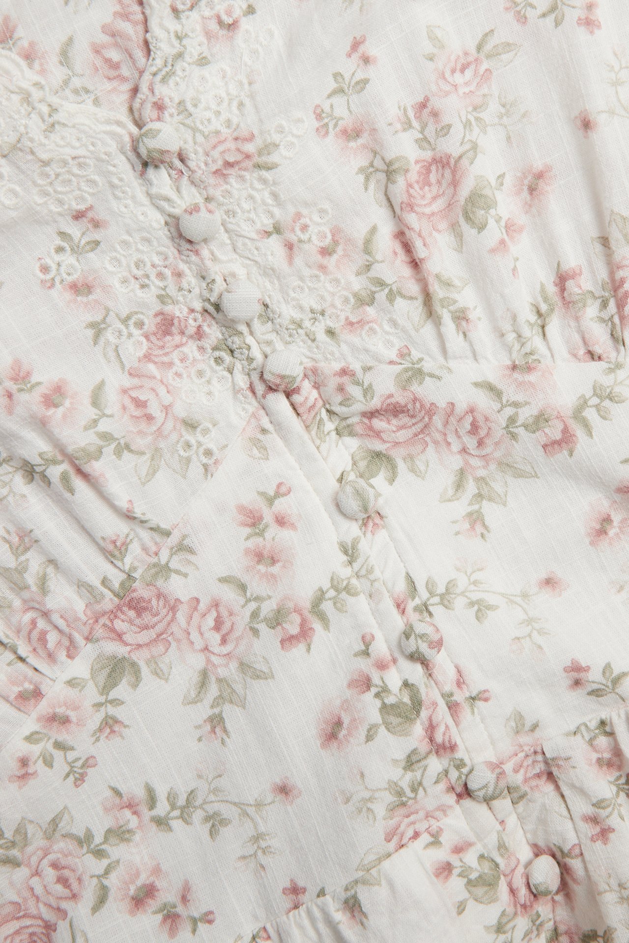 Nahaufnahme eines cremeweißen Baumwollkleides mit Blumenmuster, rosa Rosen, Stickereien und bezogenen Knöpfen.