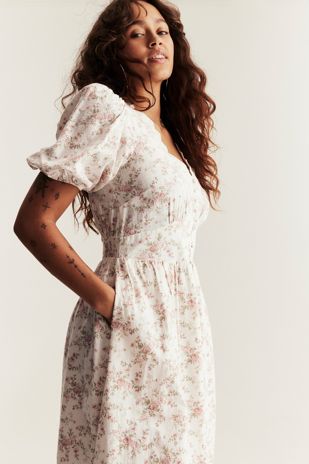 Woman in off-white floral cotton dress with puff sleeves and V-neck, hand in pocket, front-side view.