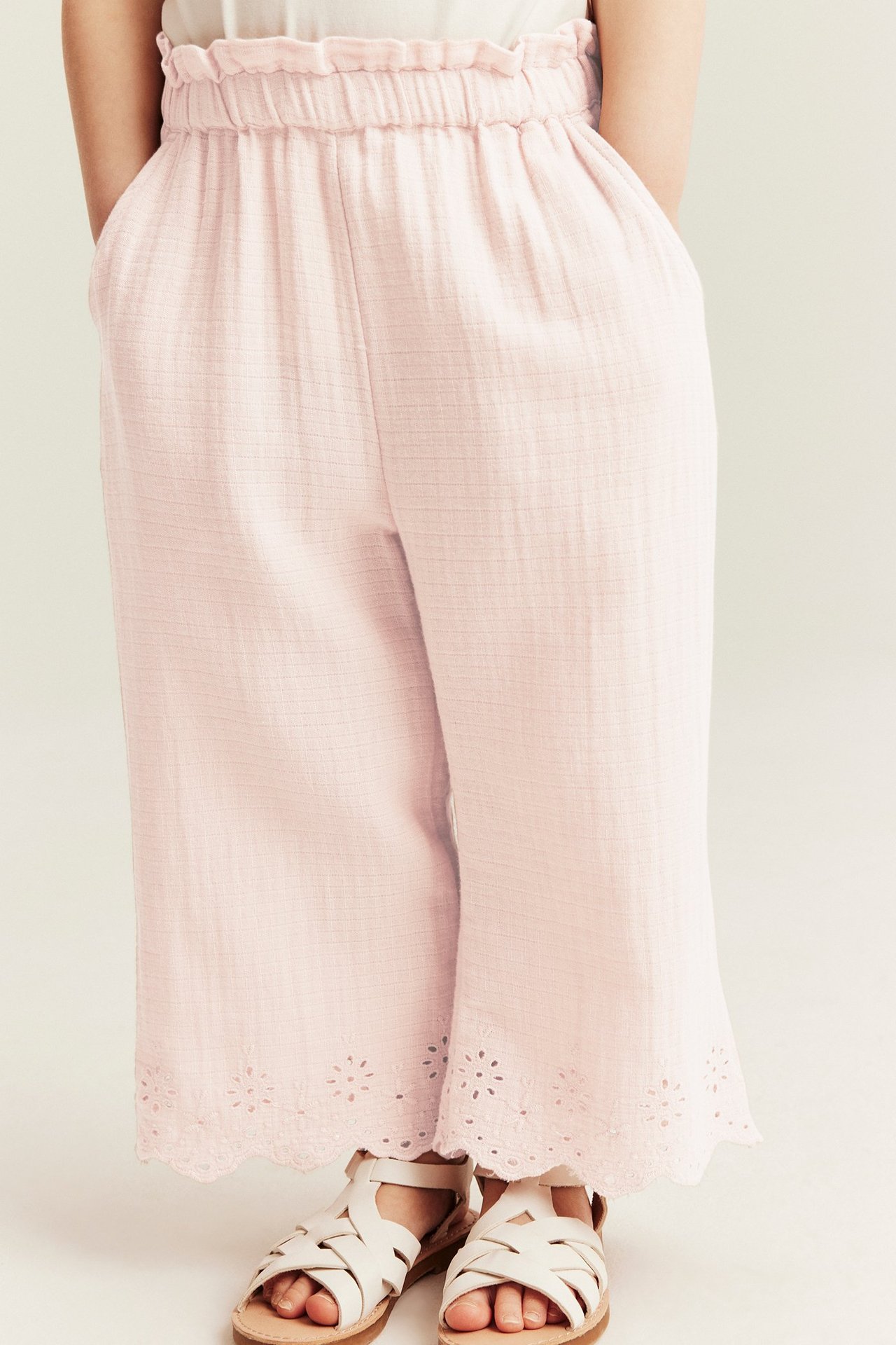 Front view of kid wearing light pink woven cotton trousers with elastic waist, embroidered hem, white sandals.