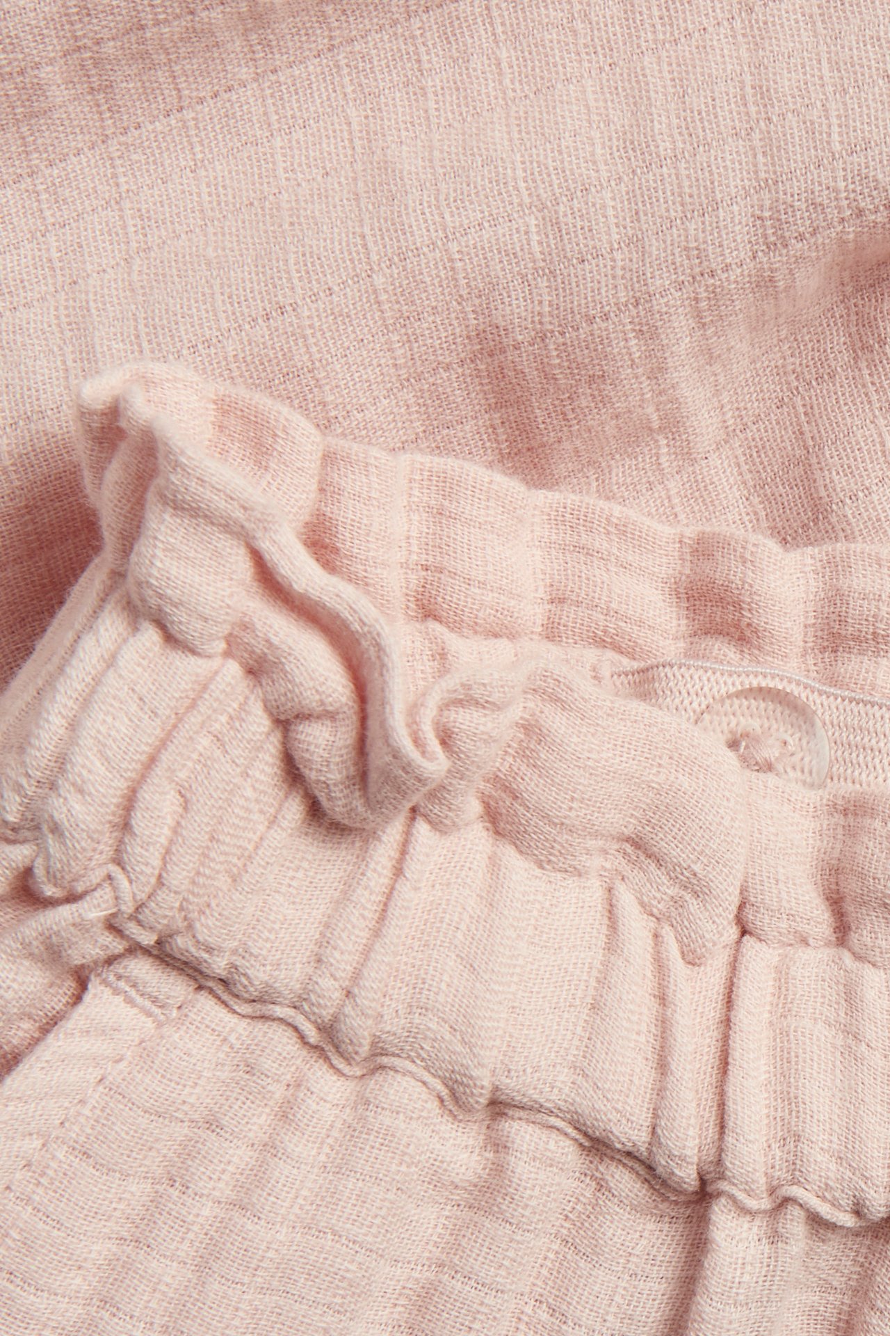 Close-up of light pink woven cotton pants with a ruffled elasticated waist and a clear button.