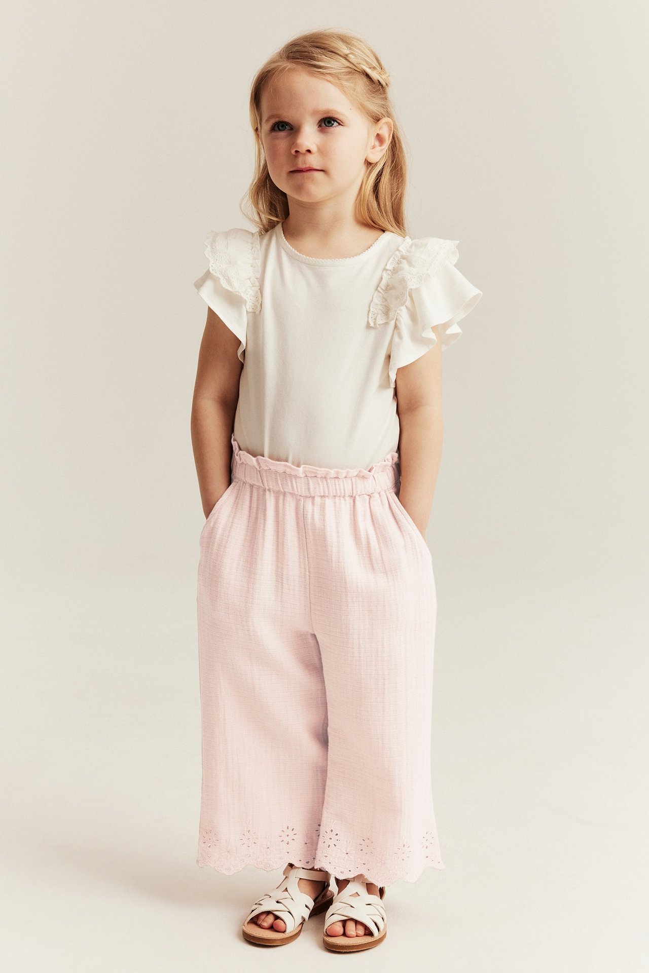 Front view: Kid in light pink woven trousers with embroidered hem, white ruffle top, and white sandals.