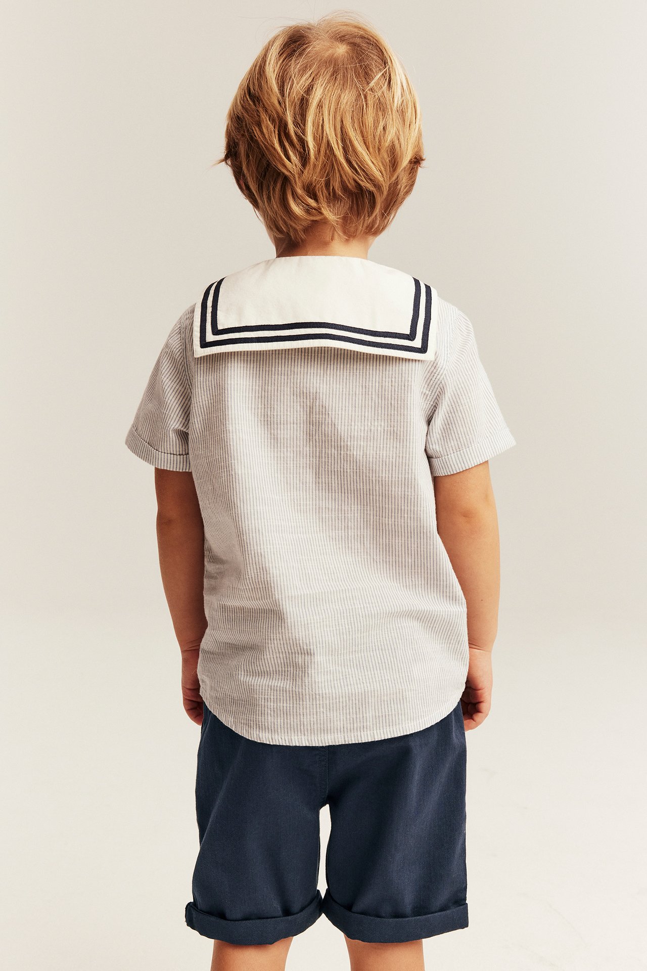 Back view of a kid in a striped blue cotton shirt with a white sailor collar and navy shorts.