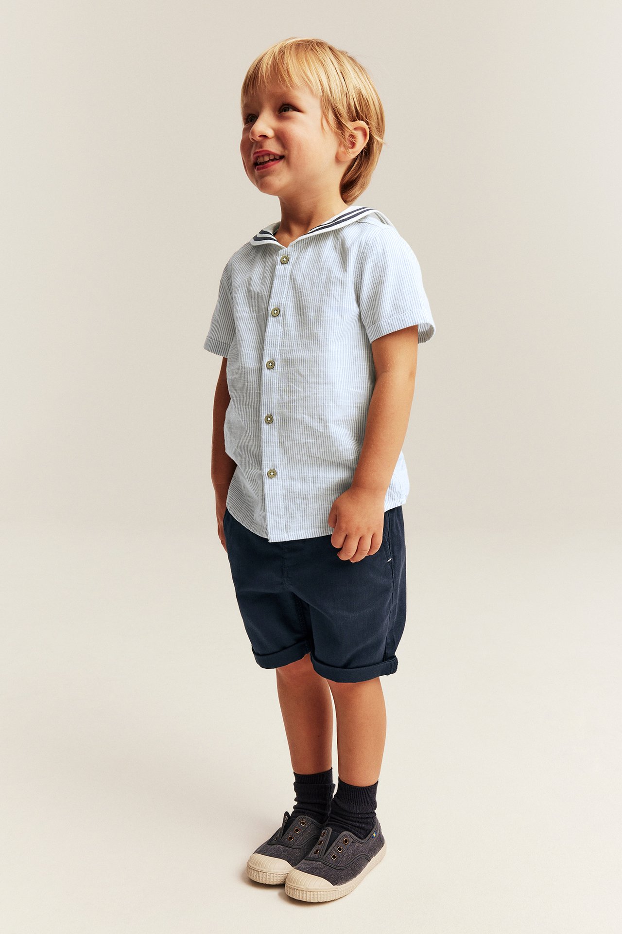 Front view: Kid in light blue striped short-sleeve sailor collar shirt, navy shorts, dark socks, and canvas shoes.