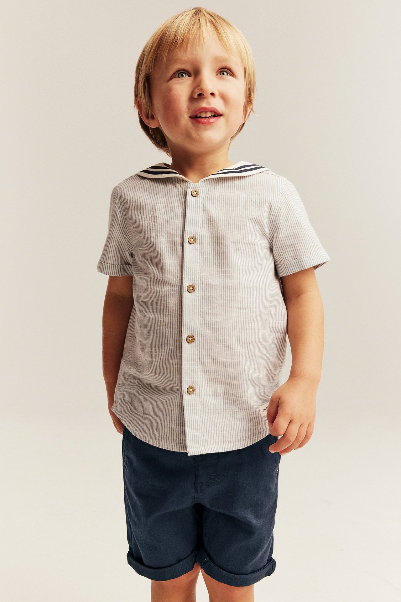 Front view of a kid wearing a blue striped short-sleeved cotton shirt with a sailor collar and navy blue shorts.