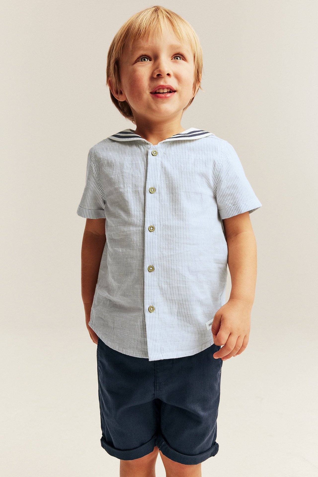 Front view: Kid in a light blue striped cotton shirt with sailor collar and dark blue shorts.