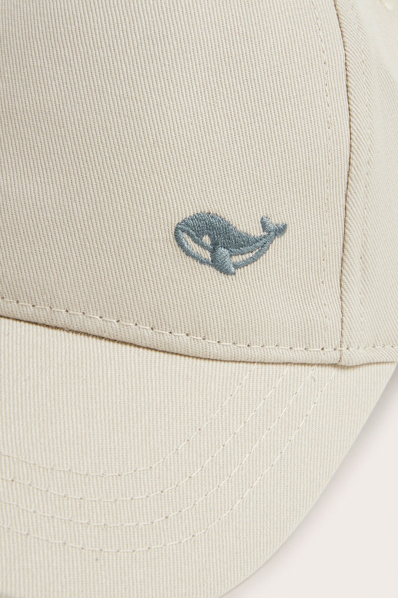 Close-up front view of a beige cotton cap with a small embroidered blue whale on the front.