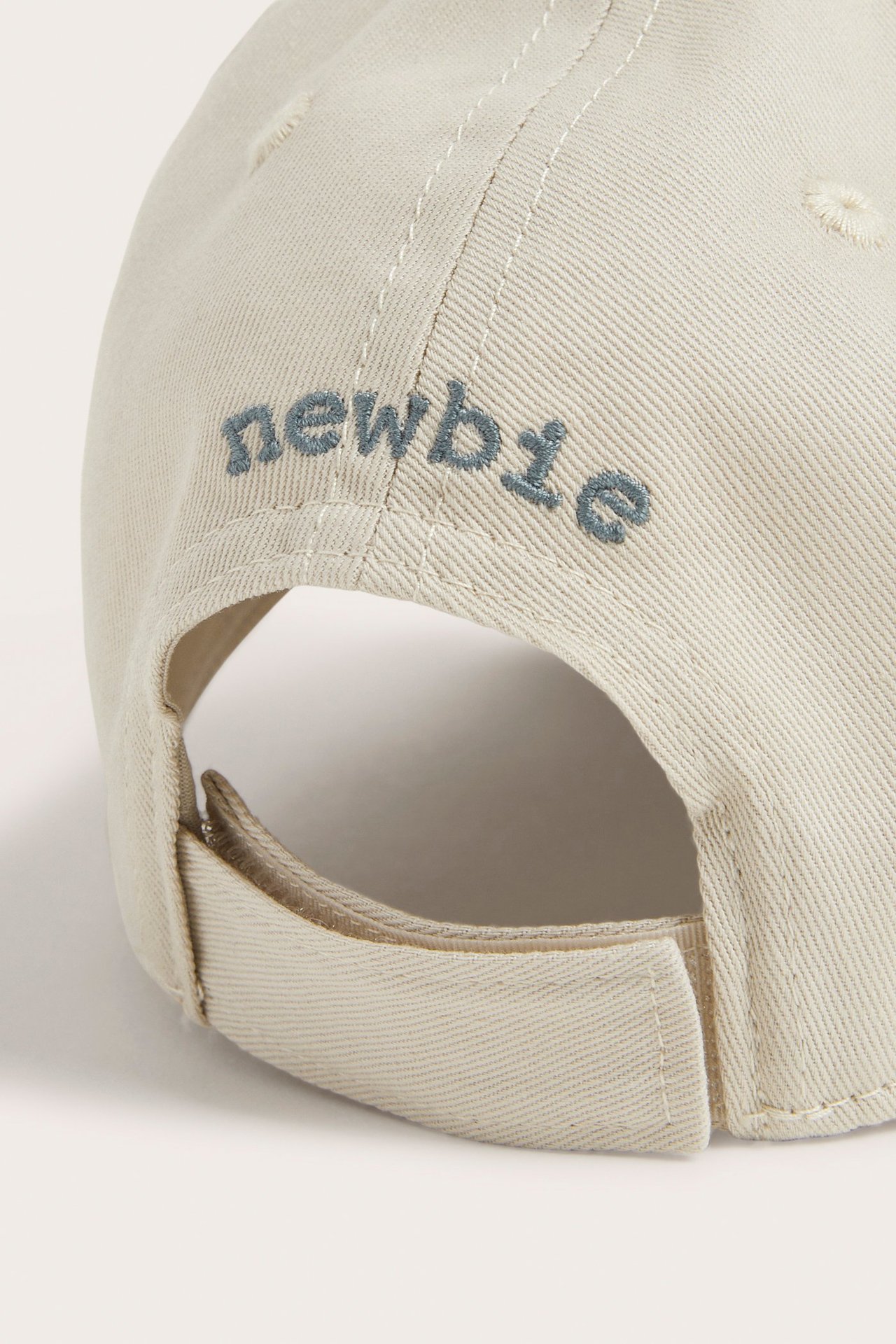 Close-up back view of a beige cap with an adjustable strap and embroidered 'newbie' text.