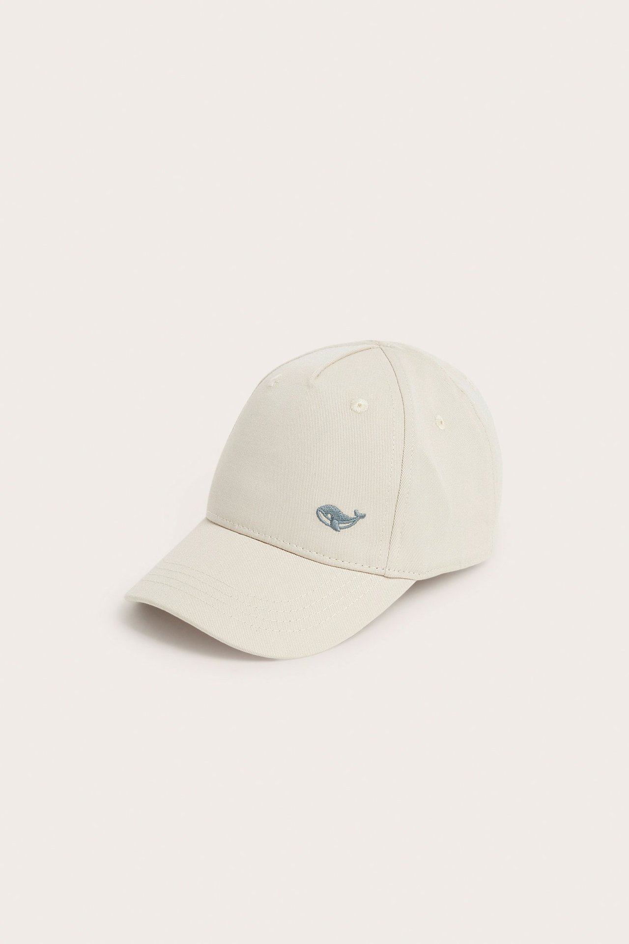 Angled front view of a beige cotton cap with a small blue whale embroidery on the front.