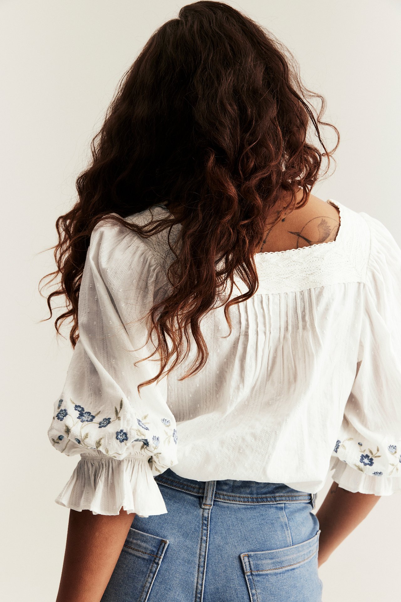 Back view of a woman in an off-white cotton blouse with embroidered ruffled cuffs and blue jeans.