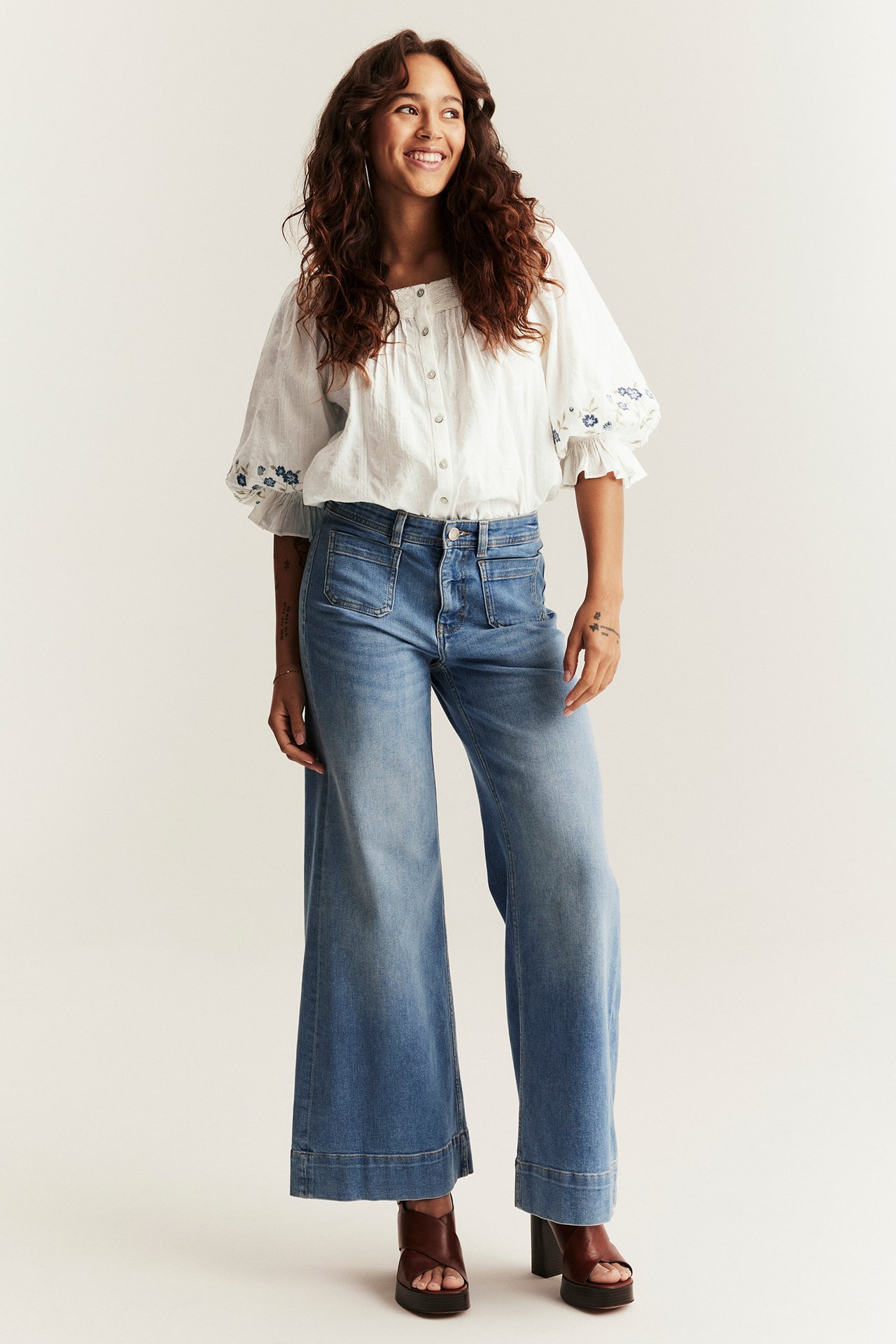 Front view of a woman wearing an offwhite cotton blouse with blue embroidery, blue wide-leg jeans, and brown platform sandals.
