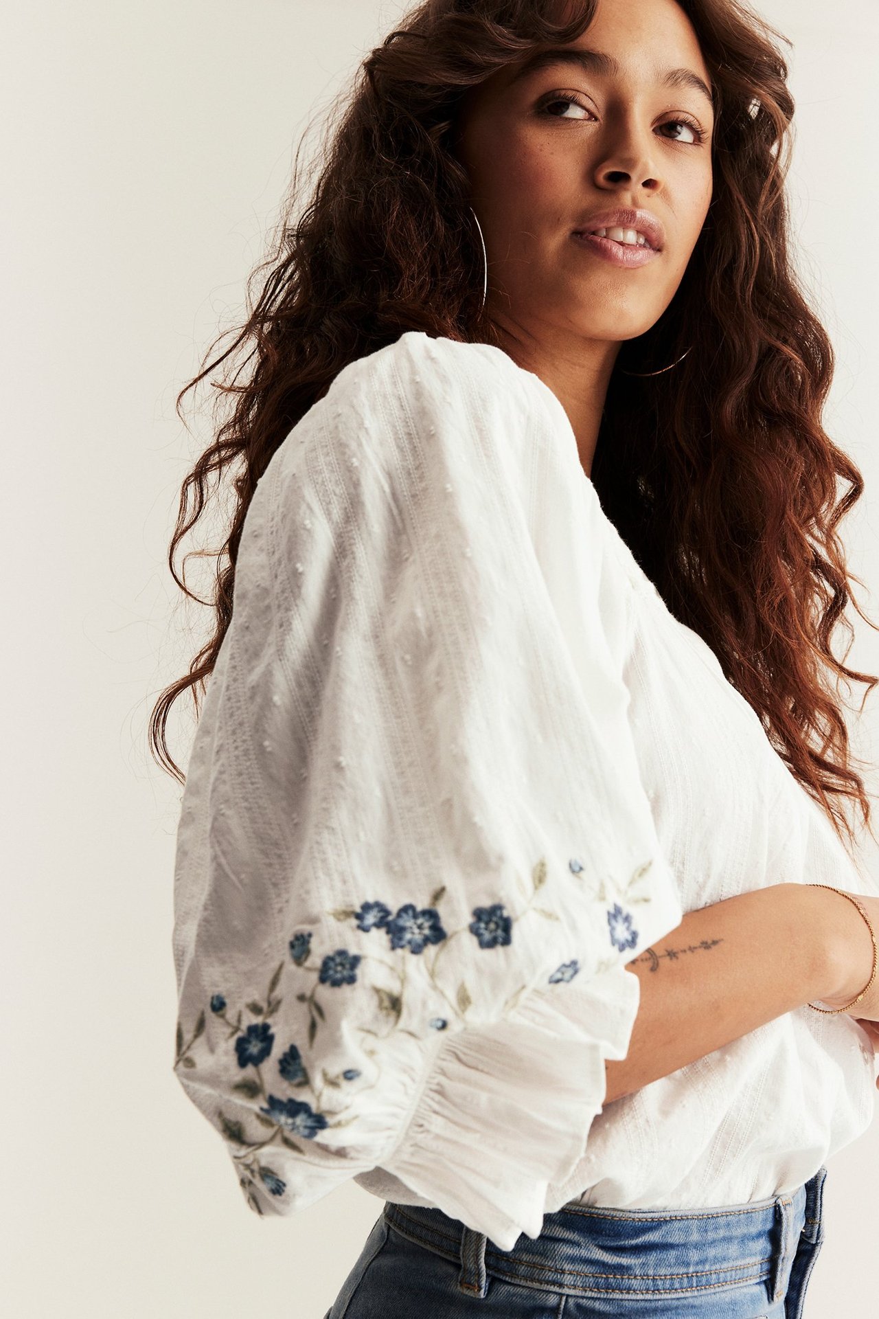 Frau trägt ecrufarbene Baumwollbluse mit blauer Blumenstickerei an Puffärmeln und blauen Jeans, Vorderansicht.