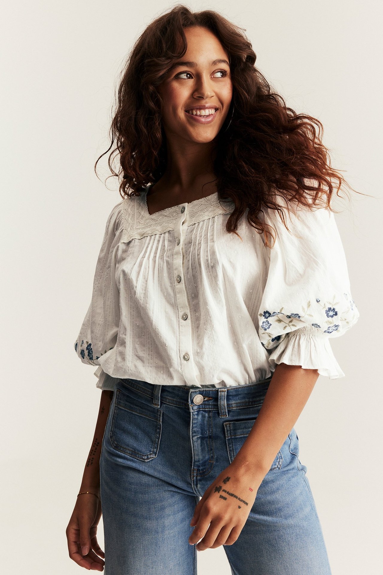 Front view of a woman in an off-white cotton blouse with lace, blue embroidered sleeves, and blue jeans.
