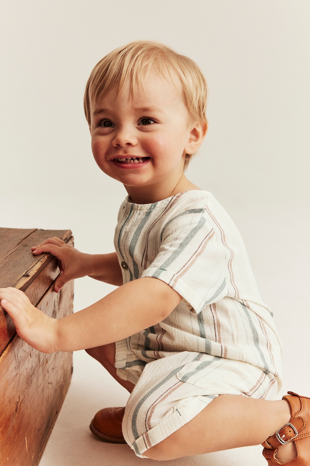 Lächelndes Baby in off-weiß gestreiftem Baumwoll-Strampler, braunen Schuhen, kauernd an Holzkiste.