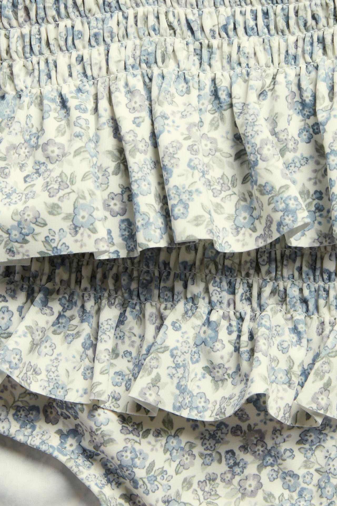 Nahaufnahme eines Kinder-Bikinis mit Smock, Rüschen und blauem Blumenmuster auf weißem Grund.