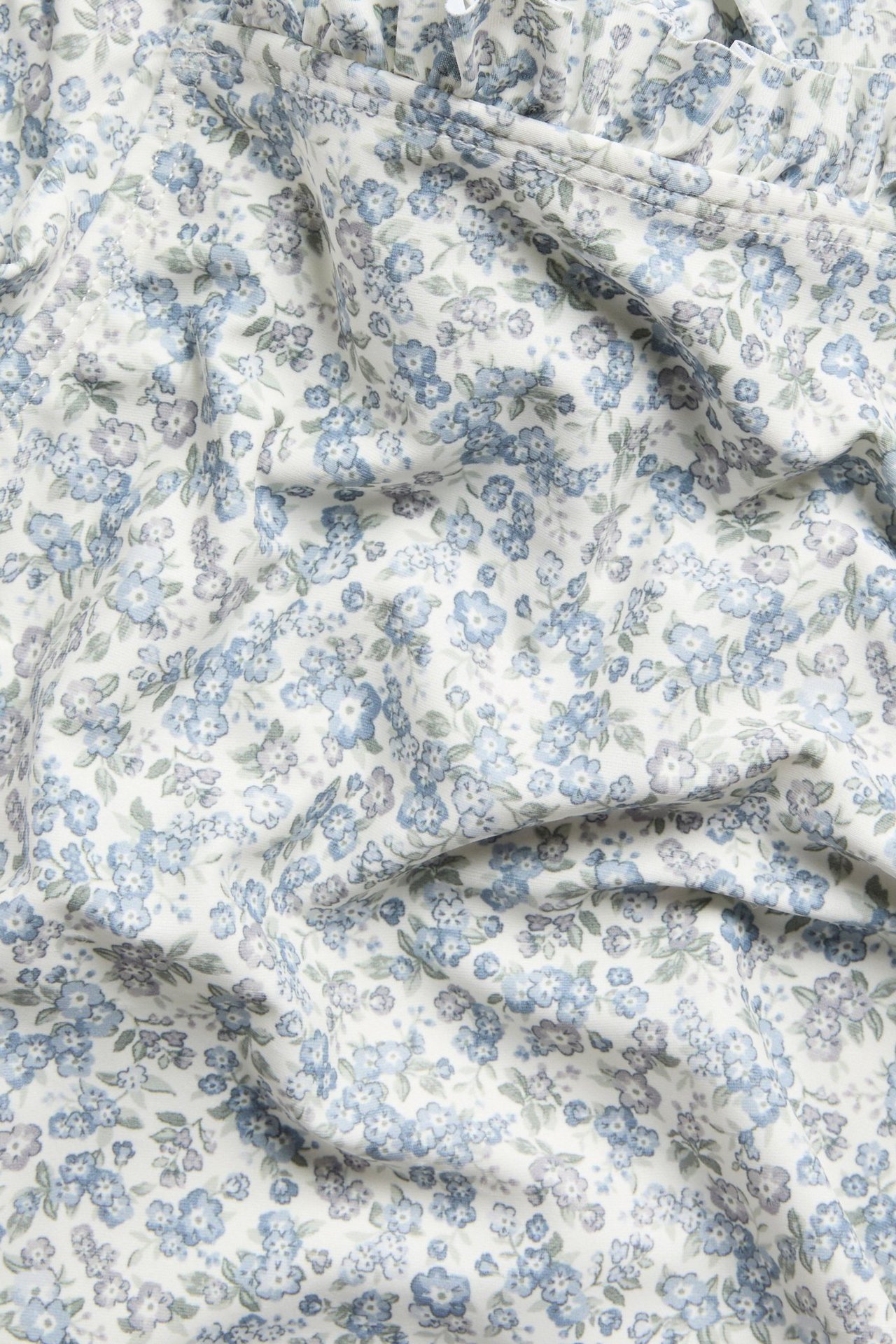 Nahaufnahme: weißer Baby-Badeanzugstoff mit blauem und hellgrünem Blumenmuster, sichtbare Rüschen.