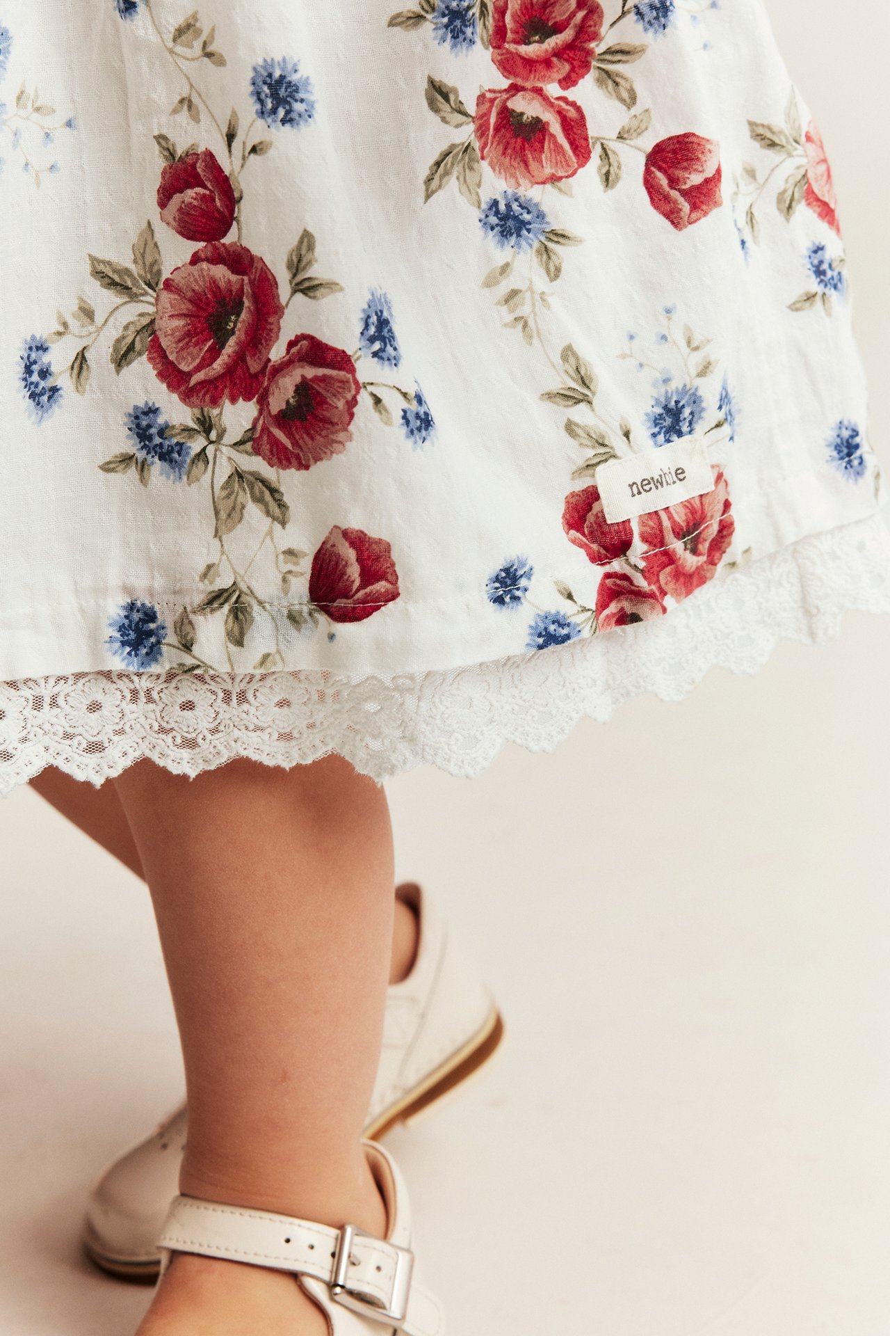 Nahaufnahme eines Kinderkleides in Off-White mit roten, blauen Blumen, Spitze, getragen mit weißen Schnallenschuhen.