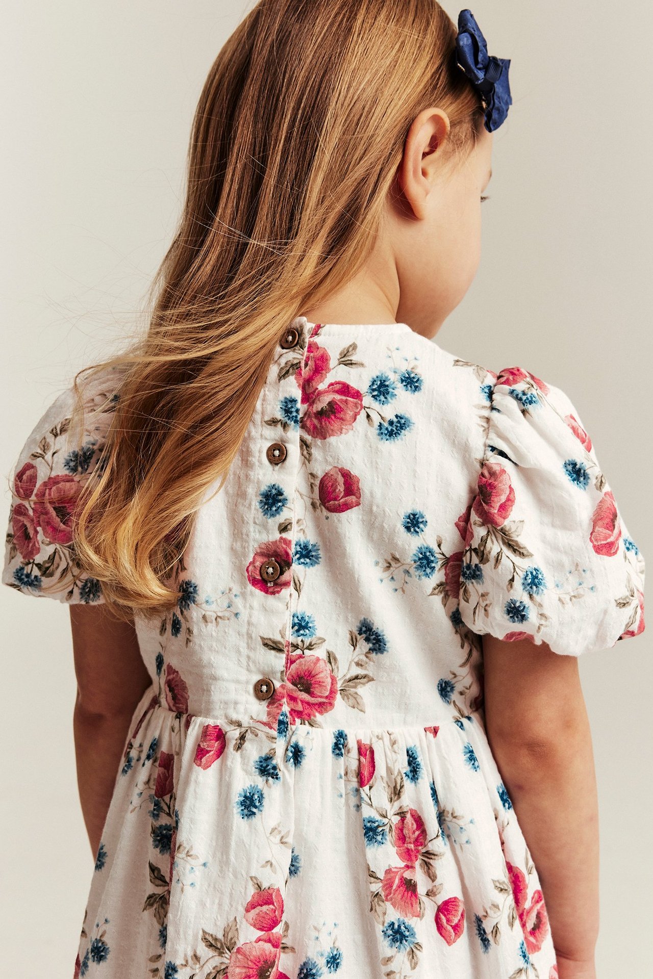 Back view of a kid in an off-white floral dress with puff sleeves, back buttons, and a blue hair bow.