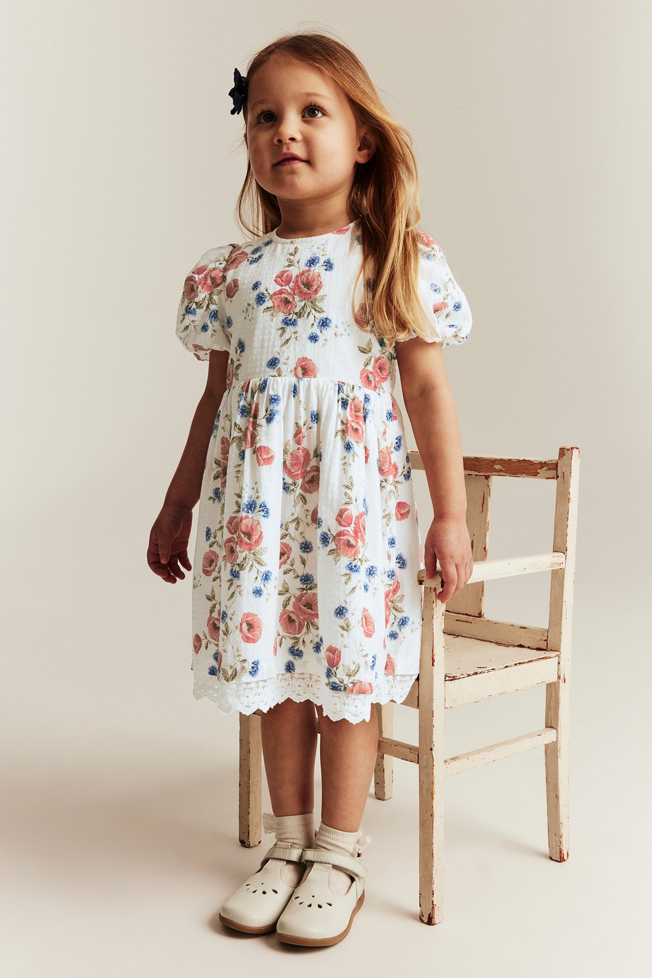 Front view: Kid wears an offwhite floral dress with puff sleeves, lace trim, and light beige shoes.
