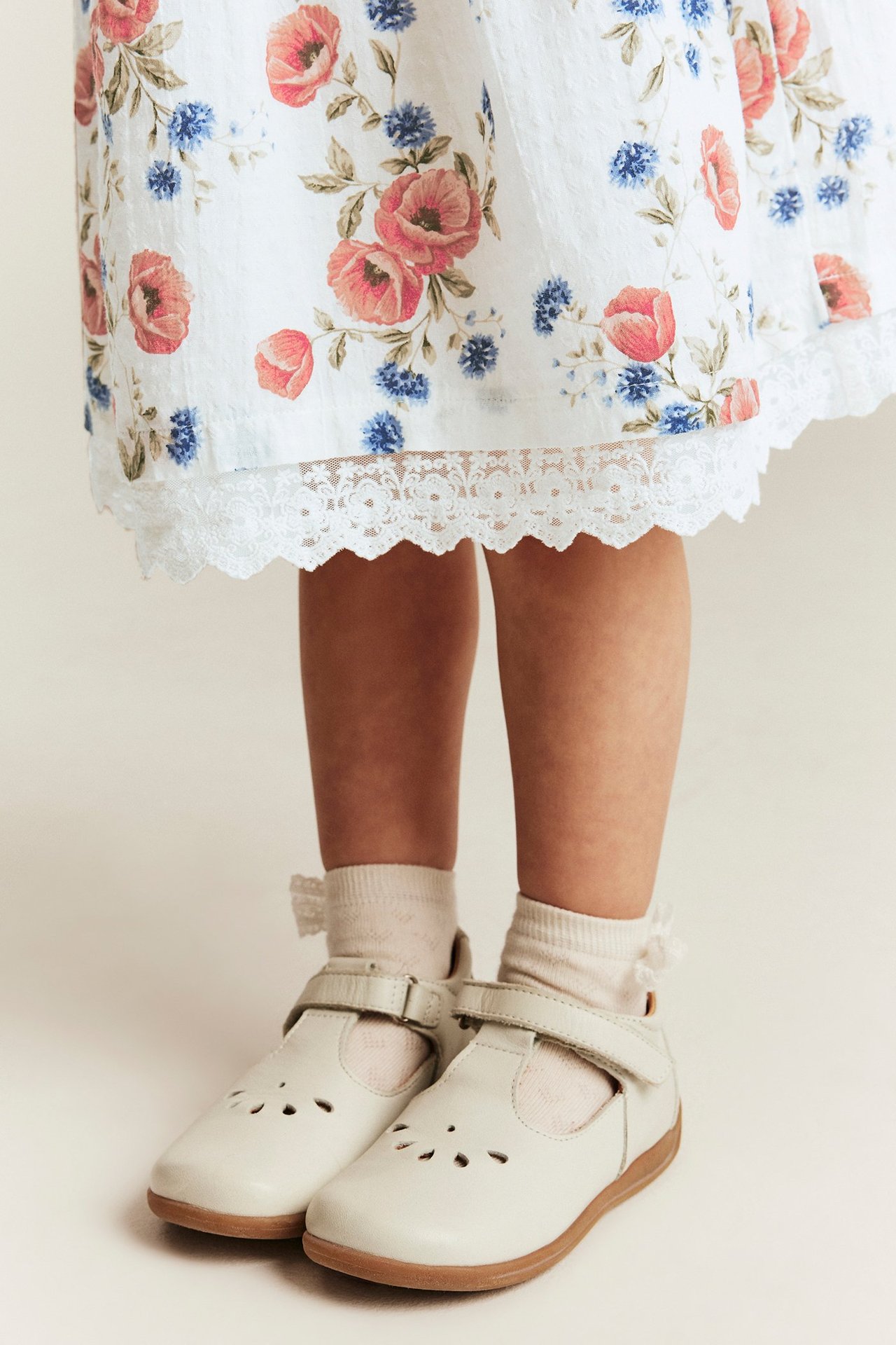 Close-up of a kid wearing an offwhite floral dress with lace hem, offwhite frilled socks, and offwhite T-bar shoes.