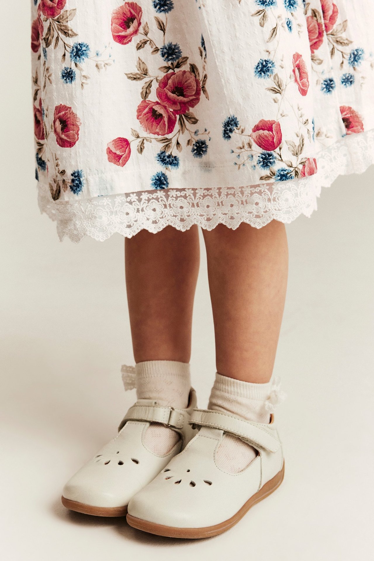 Kind im cremeweißen Blumenkleid mit Spitzenbesatz, weißen Socken und cremeweißen Mary Jane Schuhen.