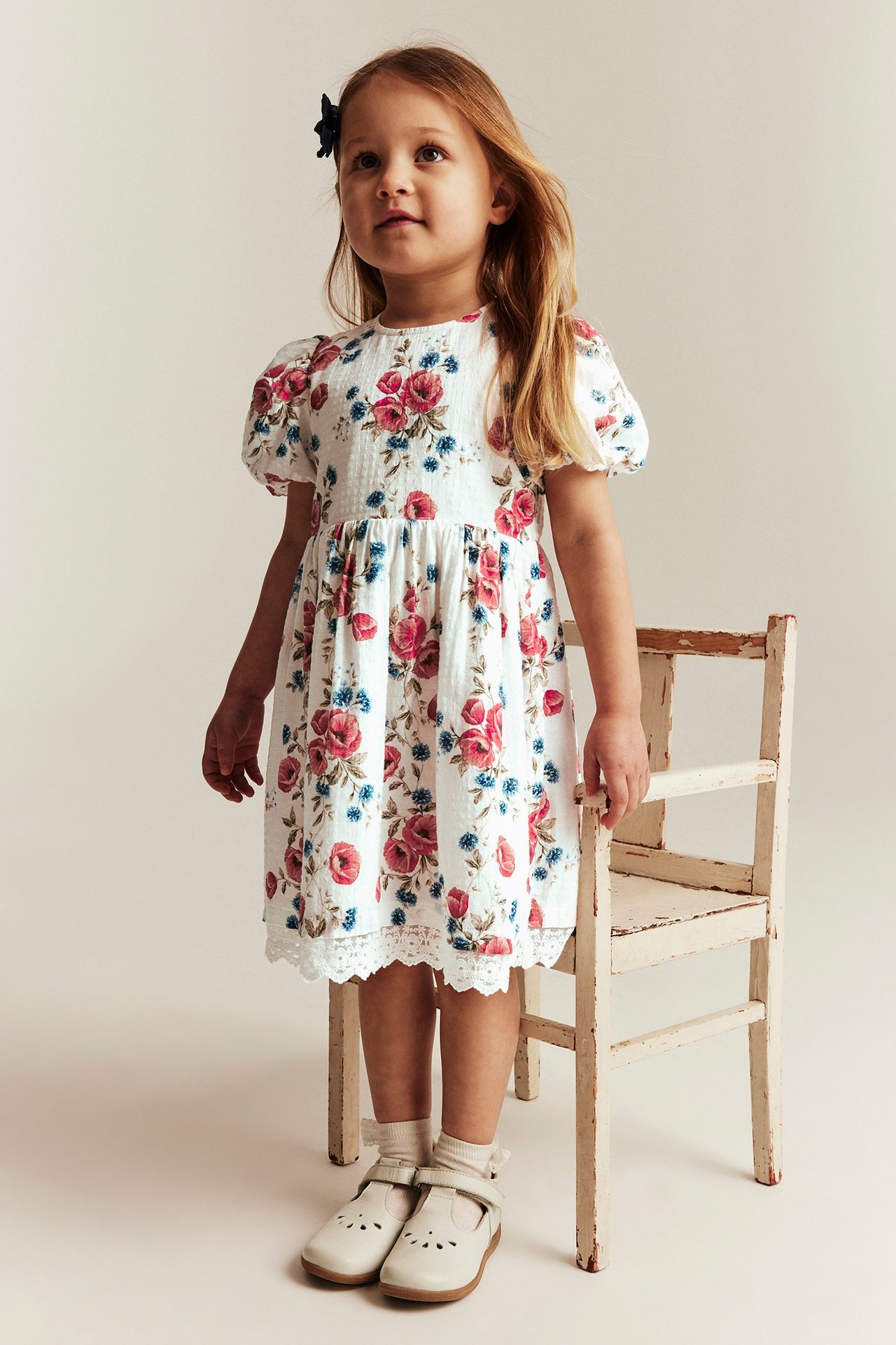 Vorderansicht eines Kindes in off-weißem Blumenkleid mit Puffärmeln, Spitzenborte und off-weißen Schuhen.