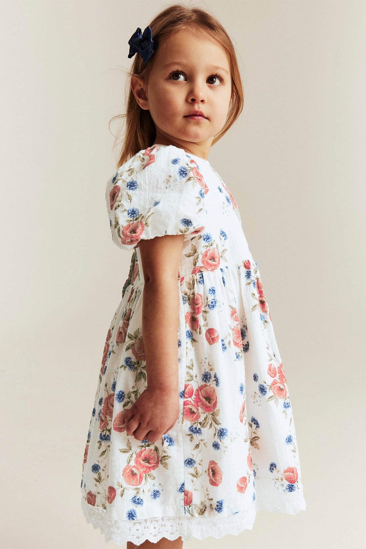 Kid in off-white floral puff-sleeved dress with lace hem and blue hair bow, side view.