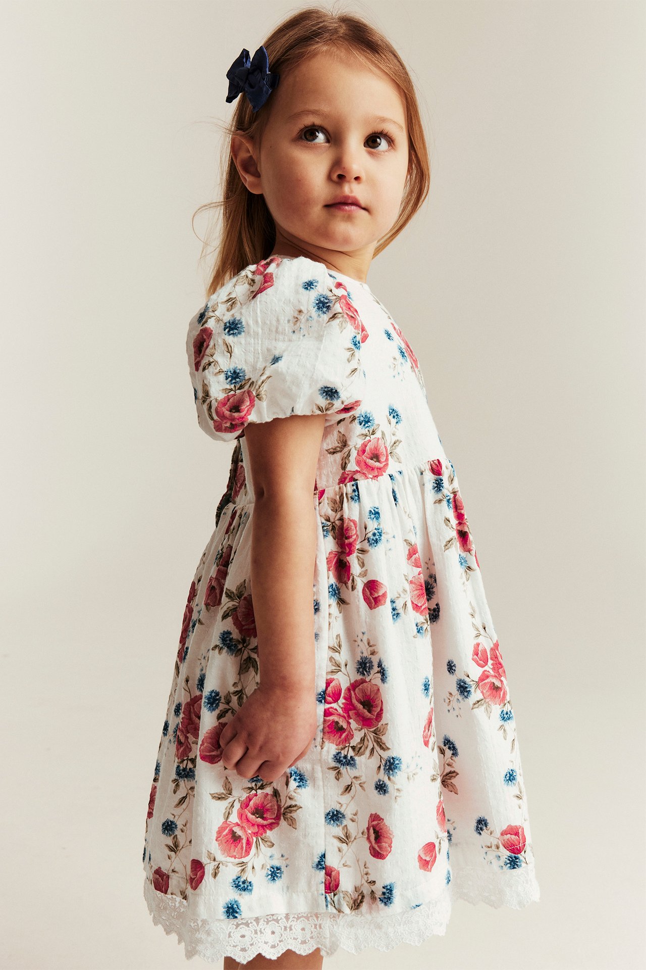 Seitenansicht eines Kindes in einem cremeweißen Blumenkleid mit Puffärmeln und einer dunkelblauen Haarspange.