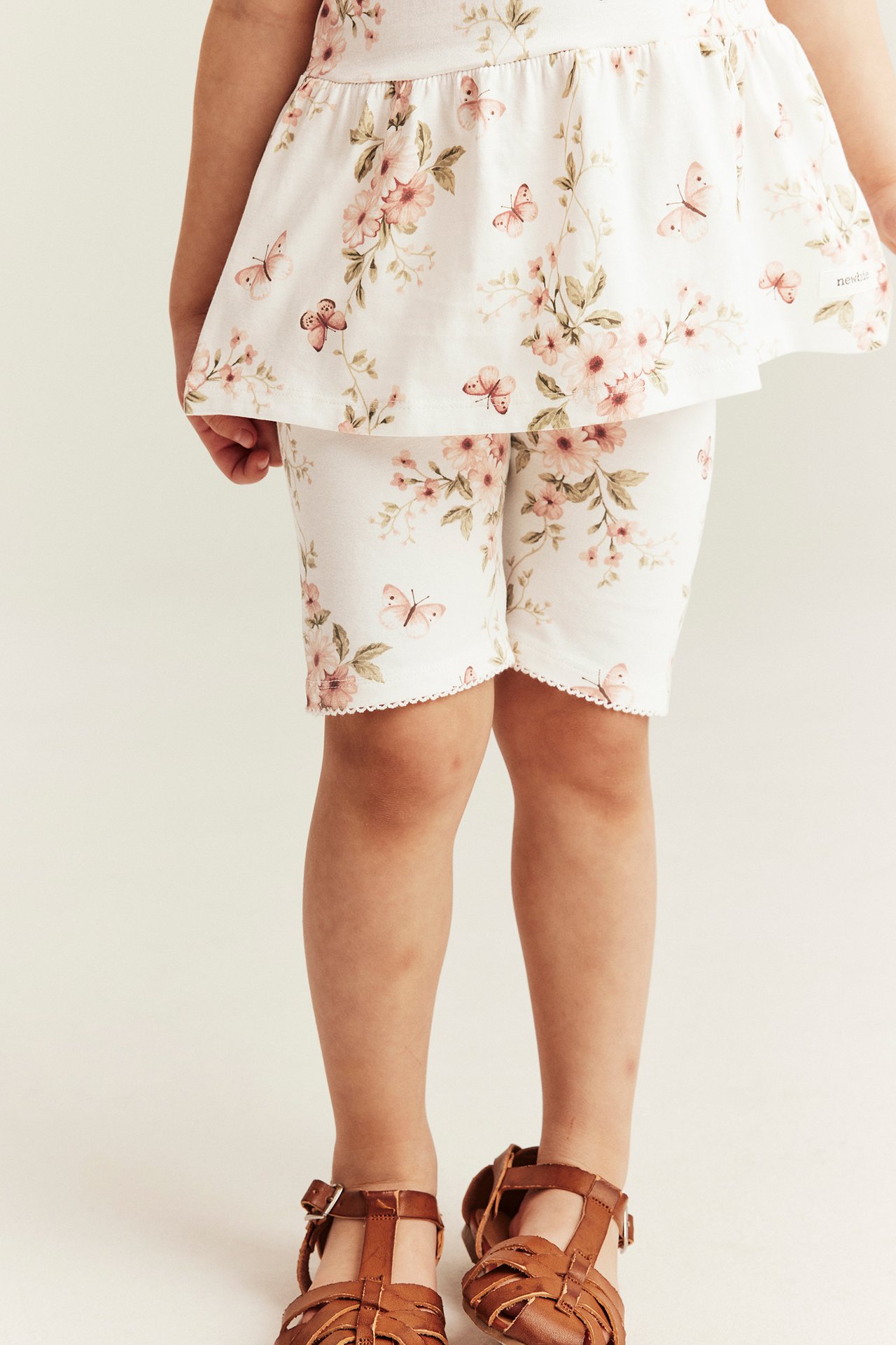 Vorderansicht: Kind trägt cremefarbene Radlerhose mit Blumenmuster und Picotkanten, passendes Oberteil und braune Sandalen.