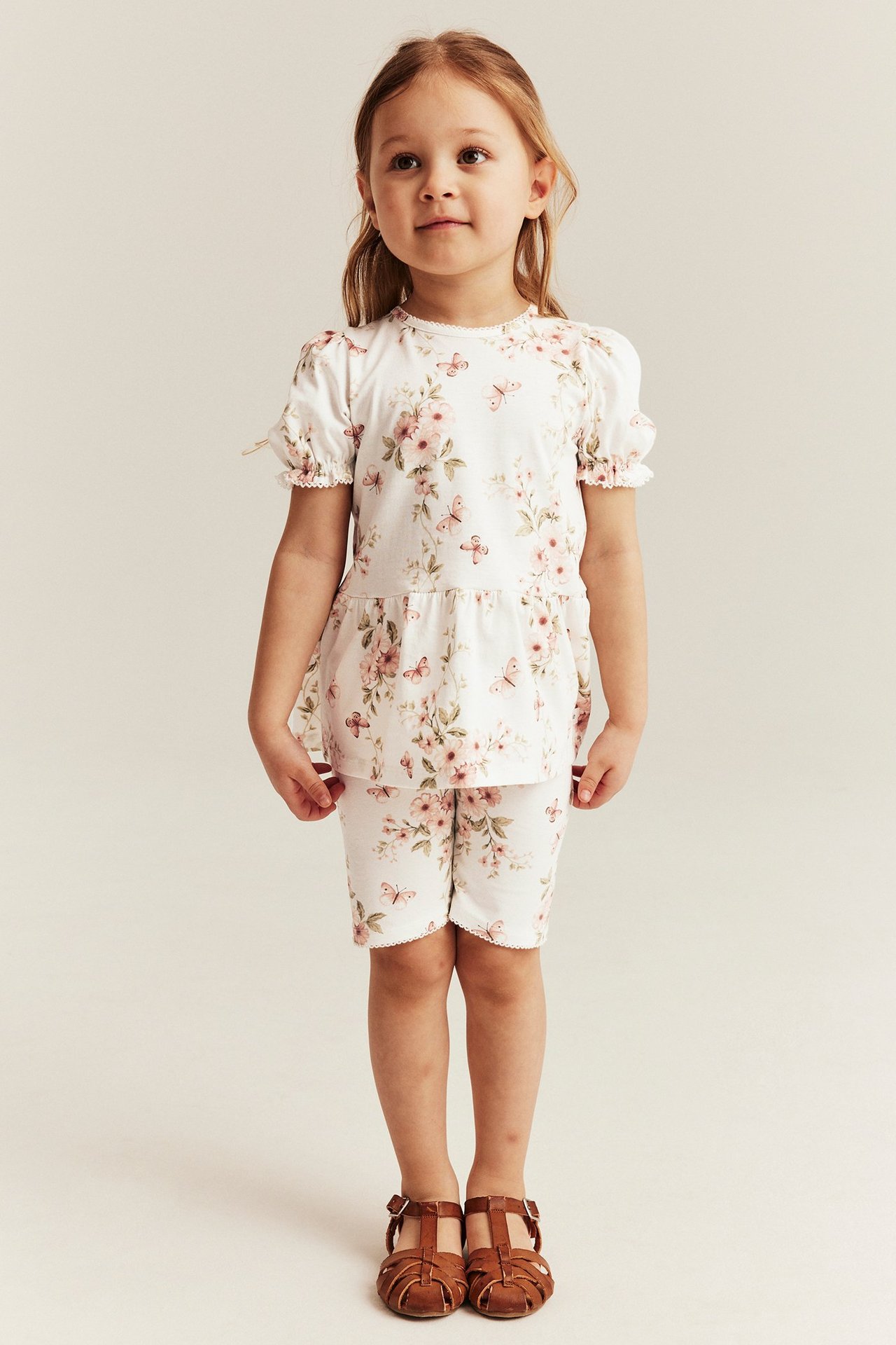 Front view of a kid in off-white floral and butterfly print bike shorts, matching top, and brown sandals.