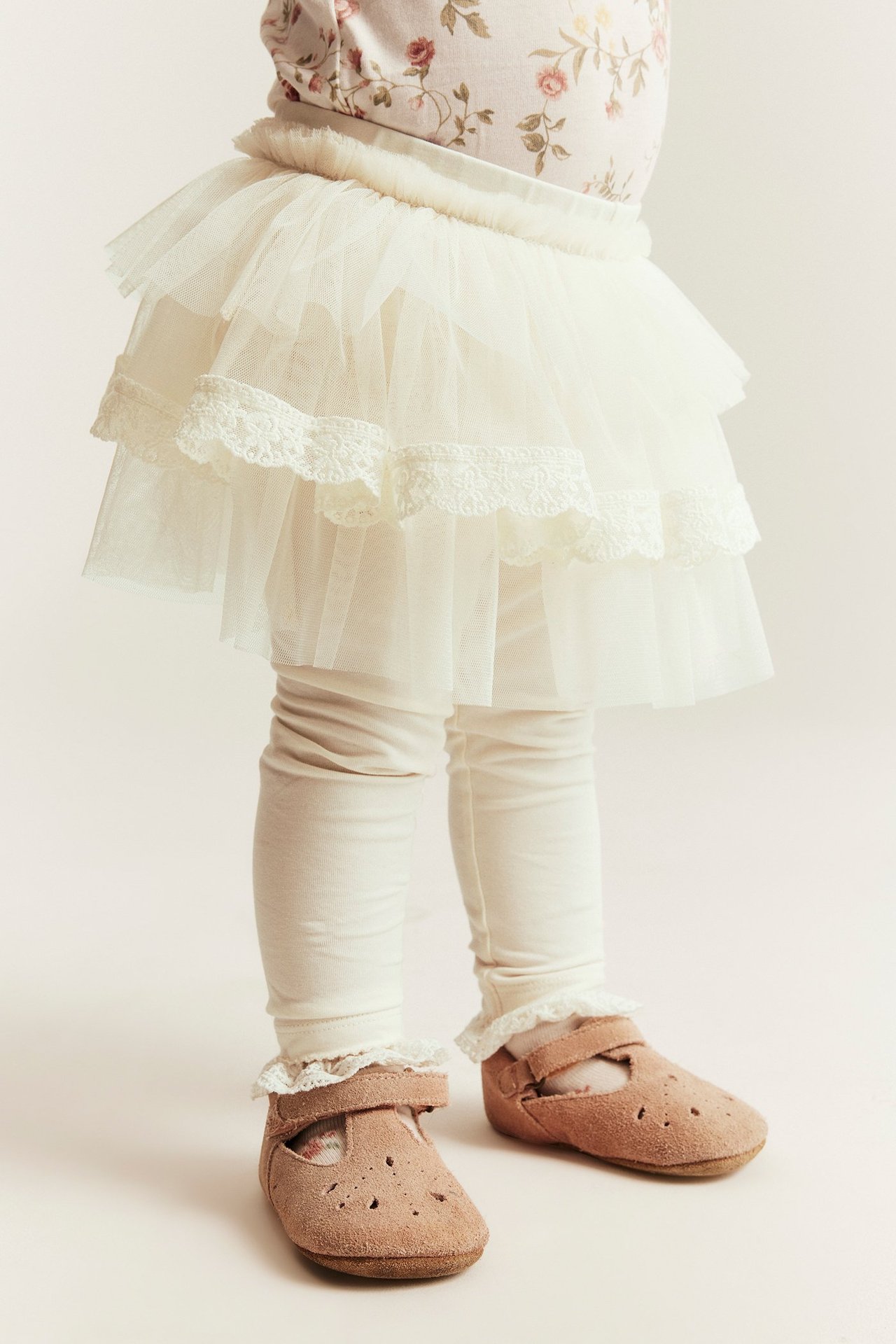 Front view: Baby wears offwhite leggings with layered tulle skirt, lace trim, and brown suede shoes.