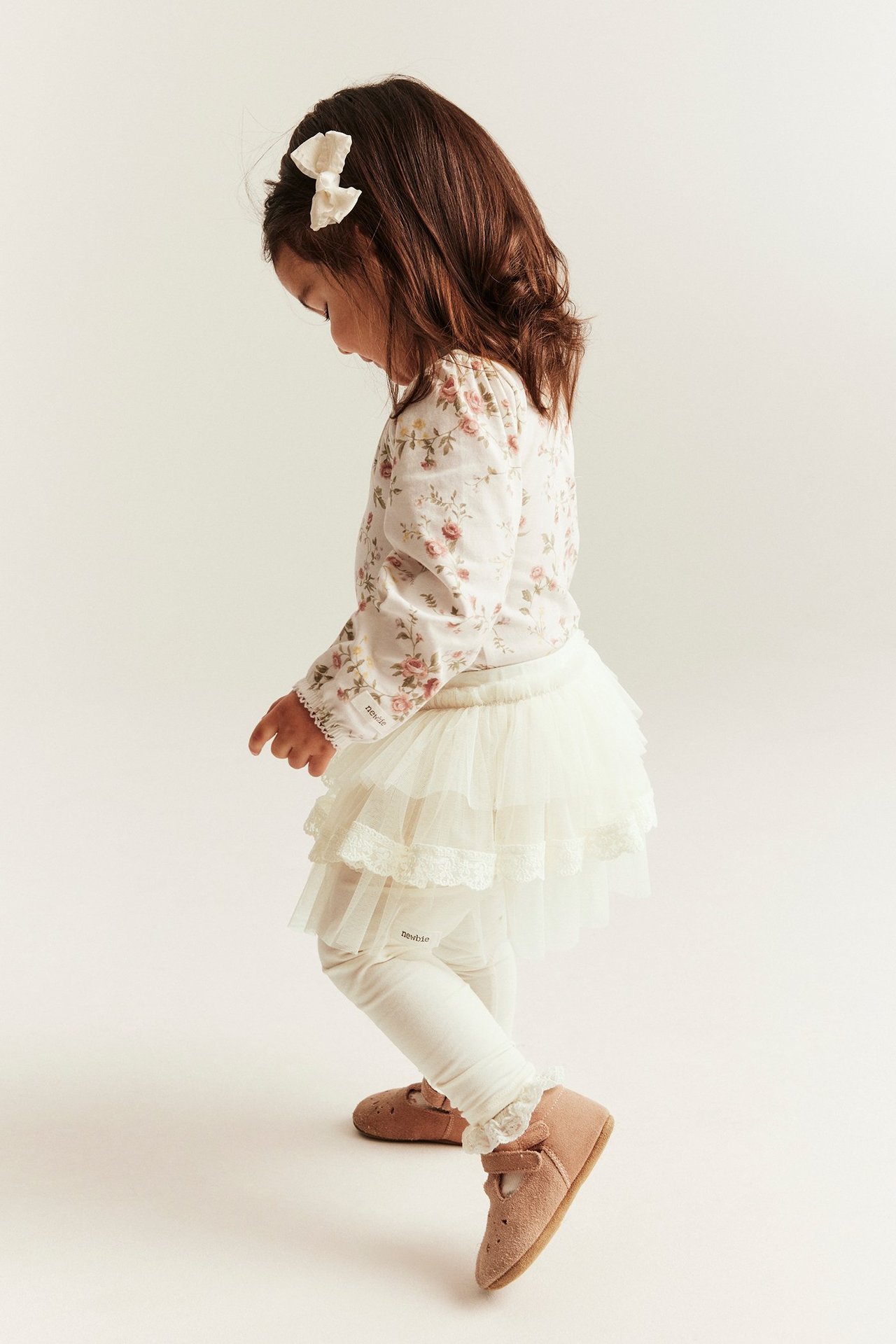 Side view of a kid wearing off-white leggings with an attached tiered tulle skirt, a floral blouse, and brown shoes.