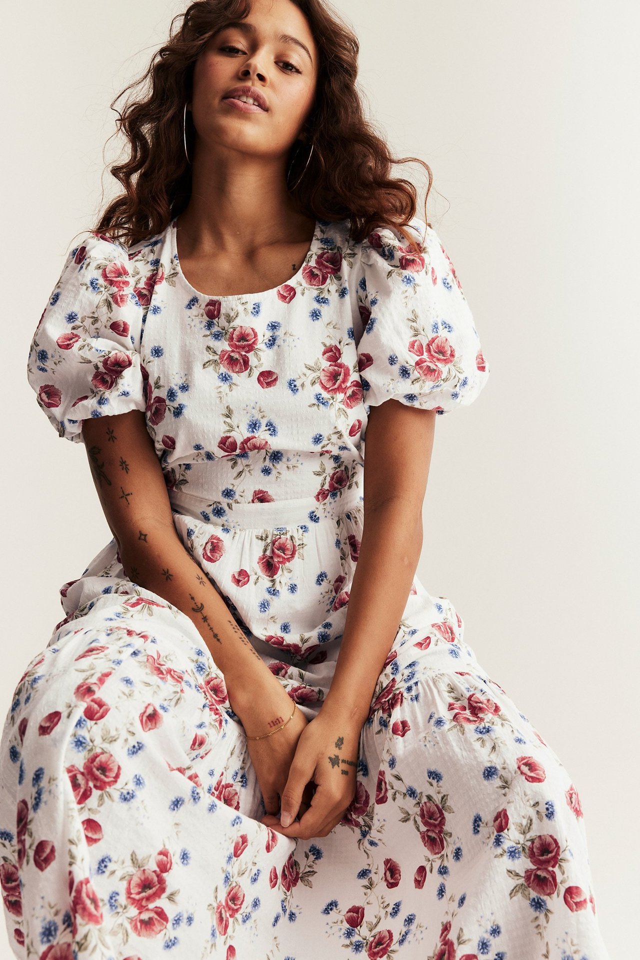 Front view of a sitting woman wearing an off-white woven floral dress with balloon sleeves and small hoop earrings.