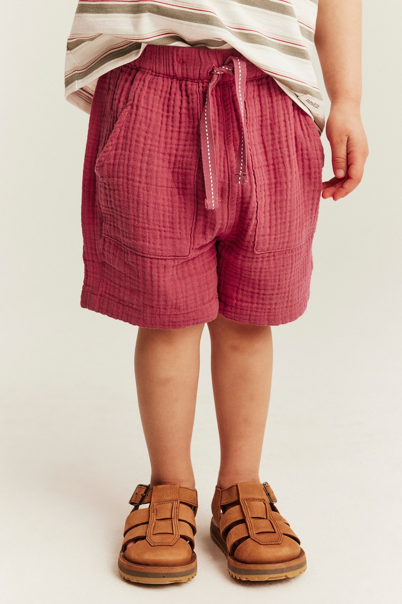 Front view, kid in red woven cotton shorts with drawstring, patch pockets, striped t-shirt, brown sandals.