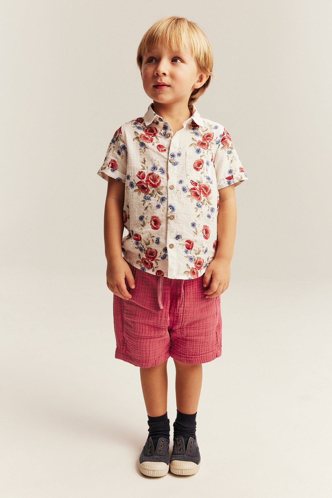Vorderansicht eines Kindes in roten Baumwollshorts, weißem Blumen-Kurzarmhemd, dunklen Socken und Leinenschuhen.