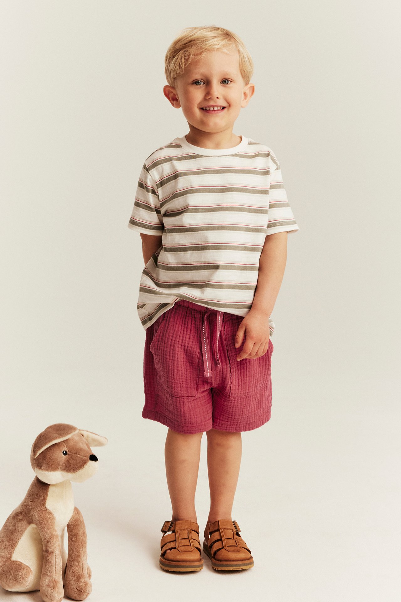Front view of a kid wearing red woven shorts with drawstring, striped t-shirt, and brown sandals.