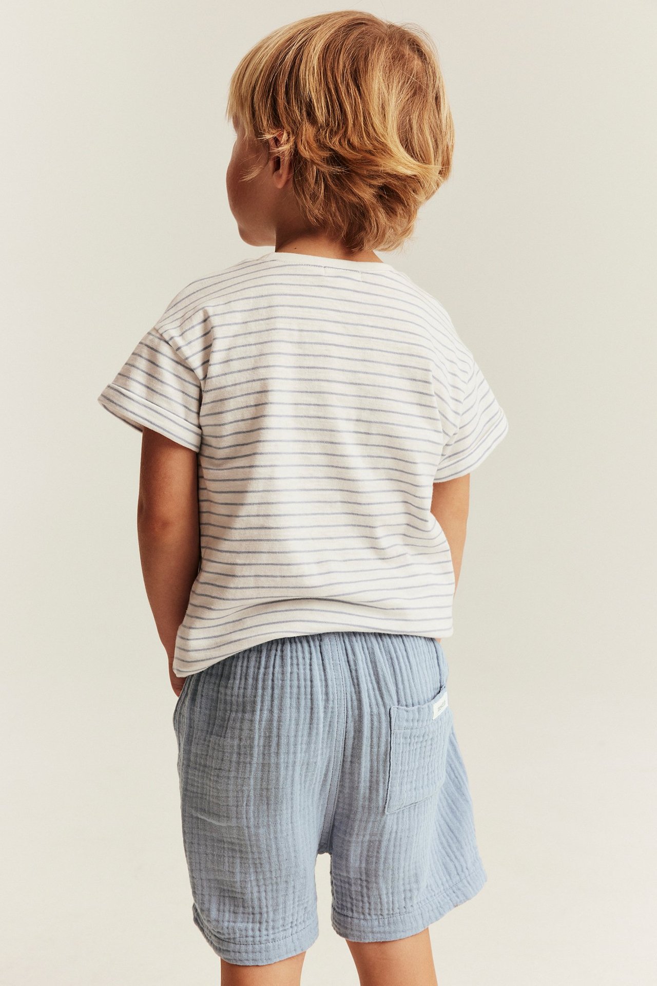 Rückansicht: Kind in blauen gewebten Baumwollshorts mit Gesäßtasche und gestreiftem T-Shirt.