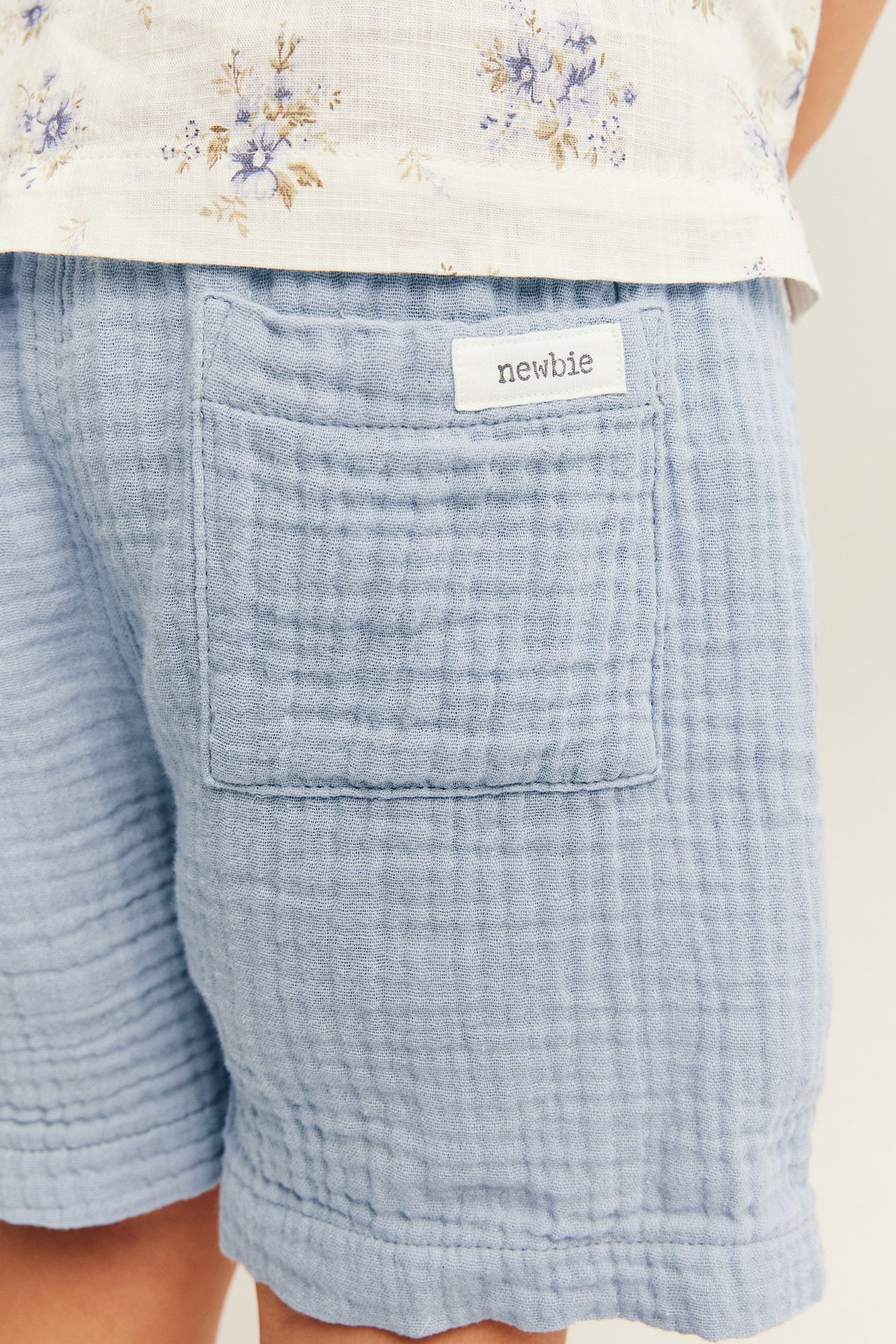Close-up back view: kid wears light blue woven shorts with a patch pocket and a white floral top.