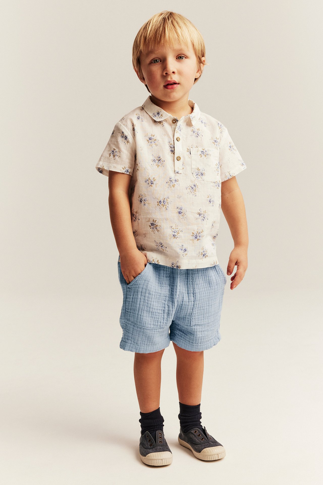 Front view of a kid wearing light blue woven cotton shorts with pockets and an off-white floral shirt.